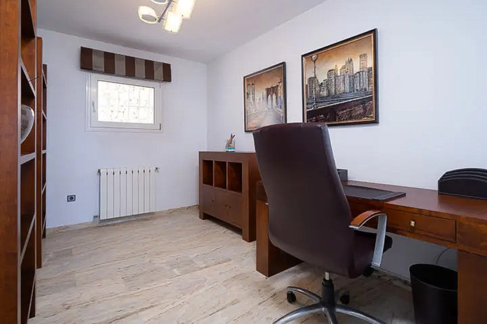 Bright home office featuring wooden furniture, a functional desk, and an ergonomic leather chair. Includes shelving, light stone flooring, and urban landscape artwork.