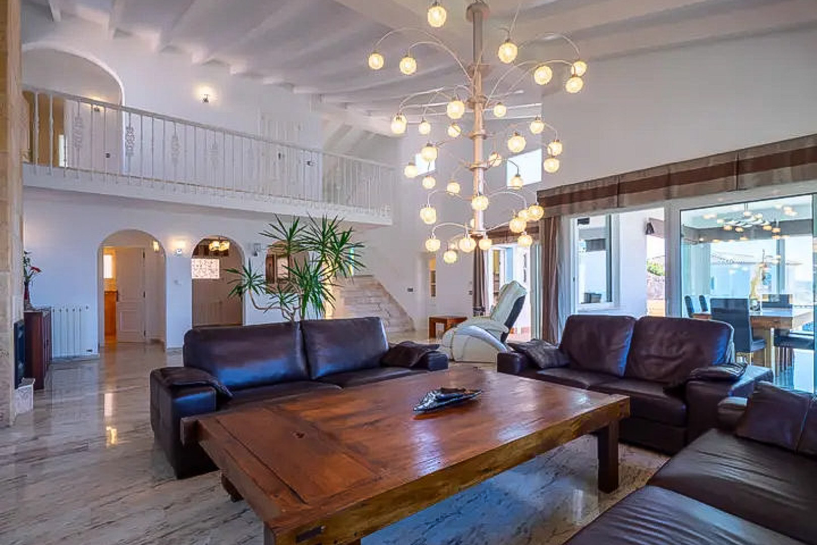 Double-height living room featuring dark leather sofas, a solid wood coffee table, and a designer chandelier. Polished marble floors and a white mezzanine gallery define the space.