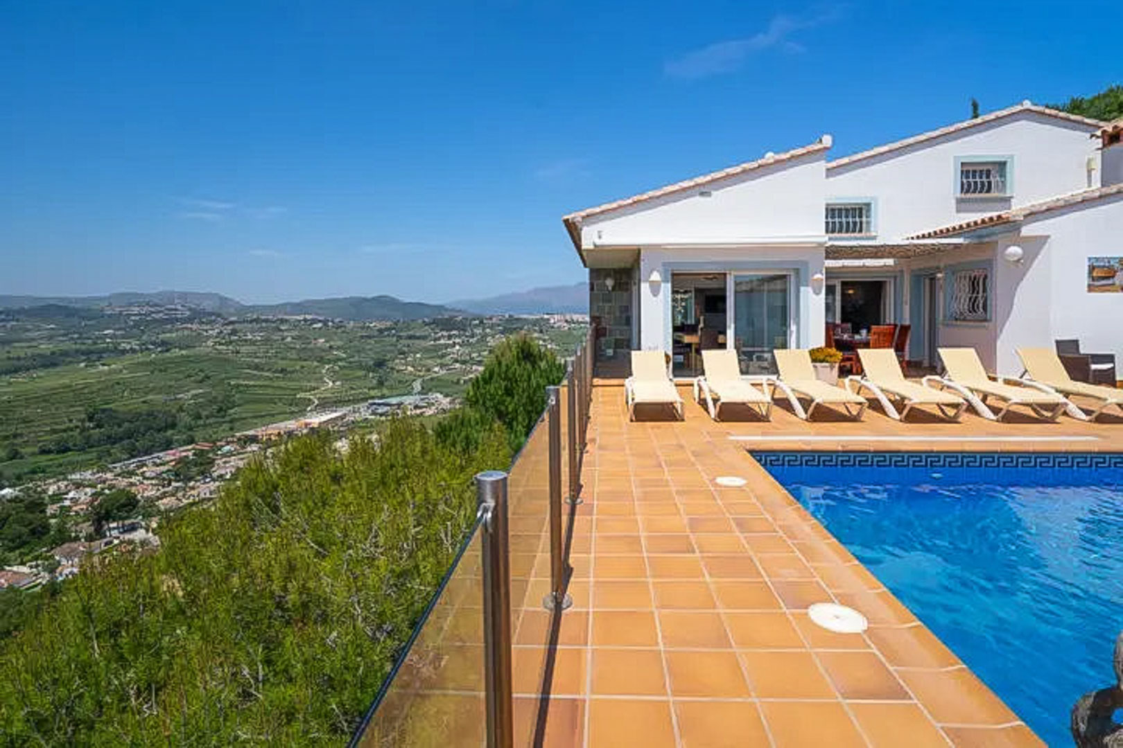 Elevated terrace featuring a blue-tiled pool, white sun loungers, and glass railings overlooking the expansive valley and mountain landscape.