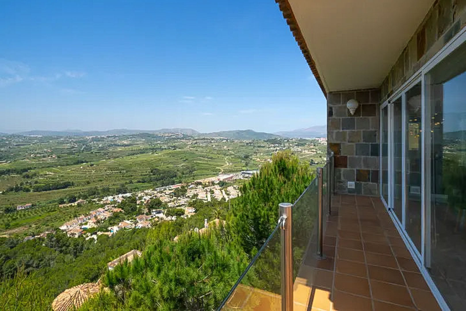 Elevated terrace featuring terracotta tiling and glass railings, overlooking the lush Moraira valley and distant mountain ranges.