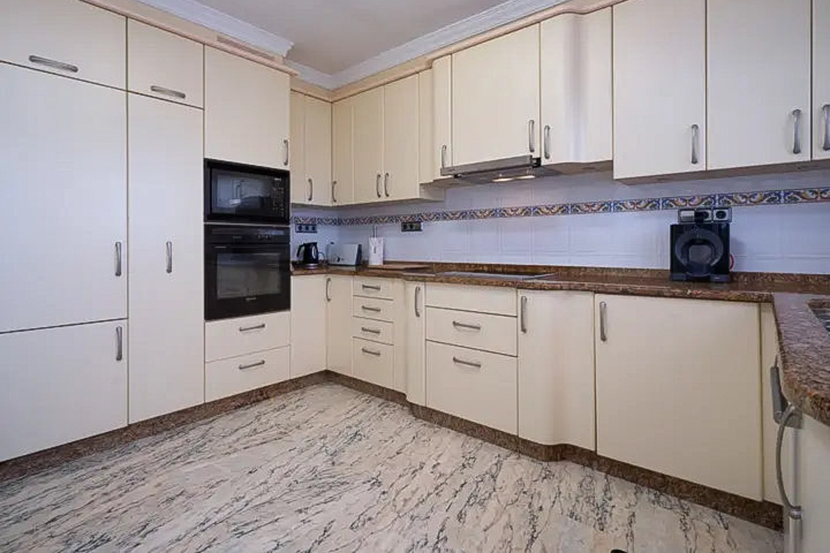 Fully equipped kitchen featuring cream cabinetry, brown granite countertops, and integrated appliances. The space is finished with polished marble flooring and a decorative tile backsplash.