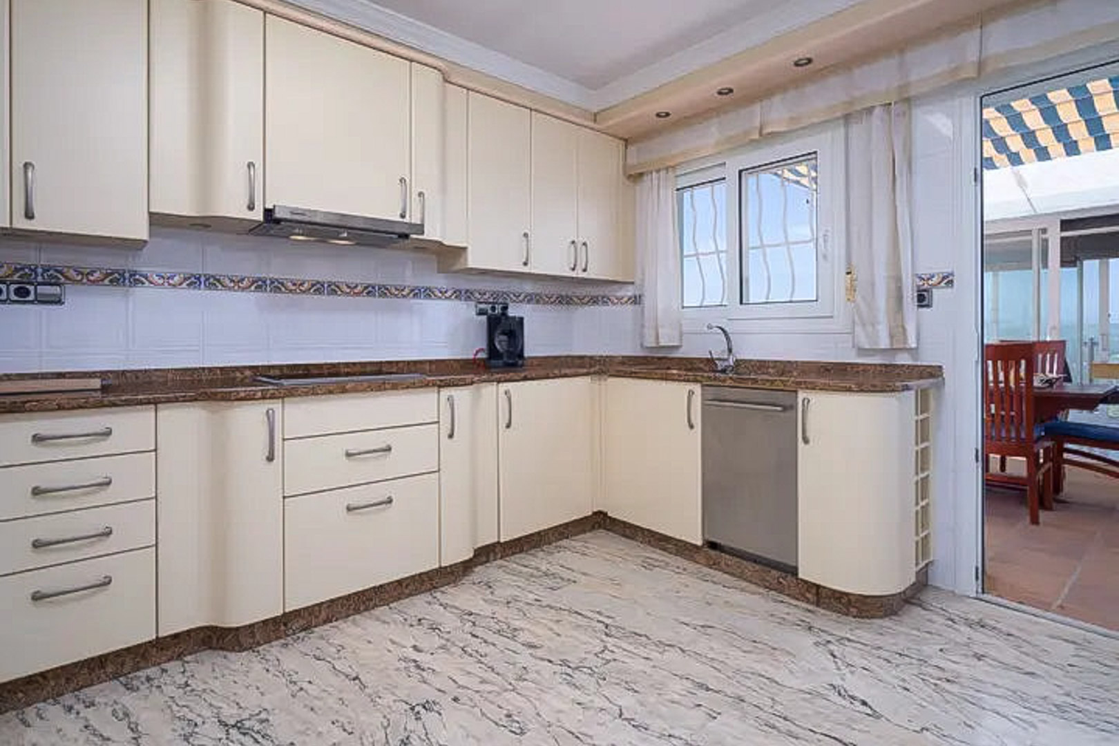 Fully equipped L-shaped kitchen with cream cabinetry, brown granite countertops, and marble floors. A glass door connects the space to the outdoor terrace.