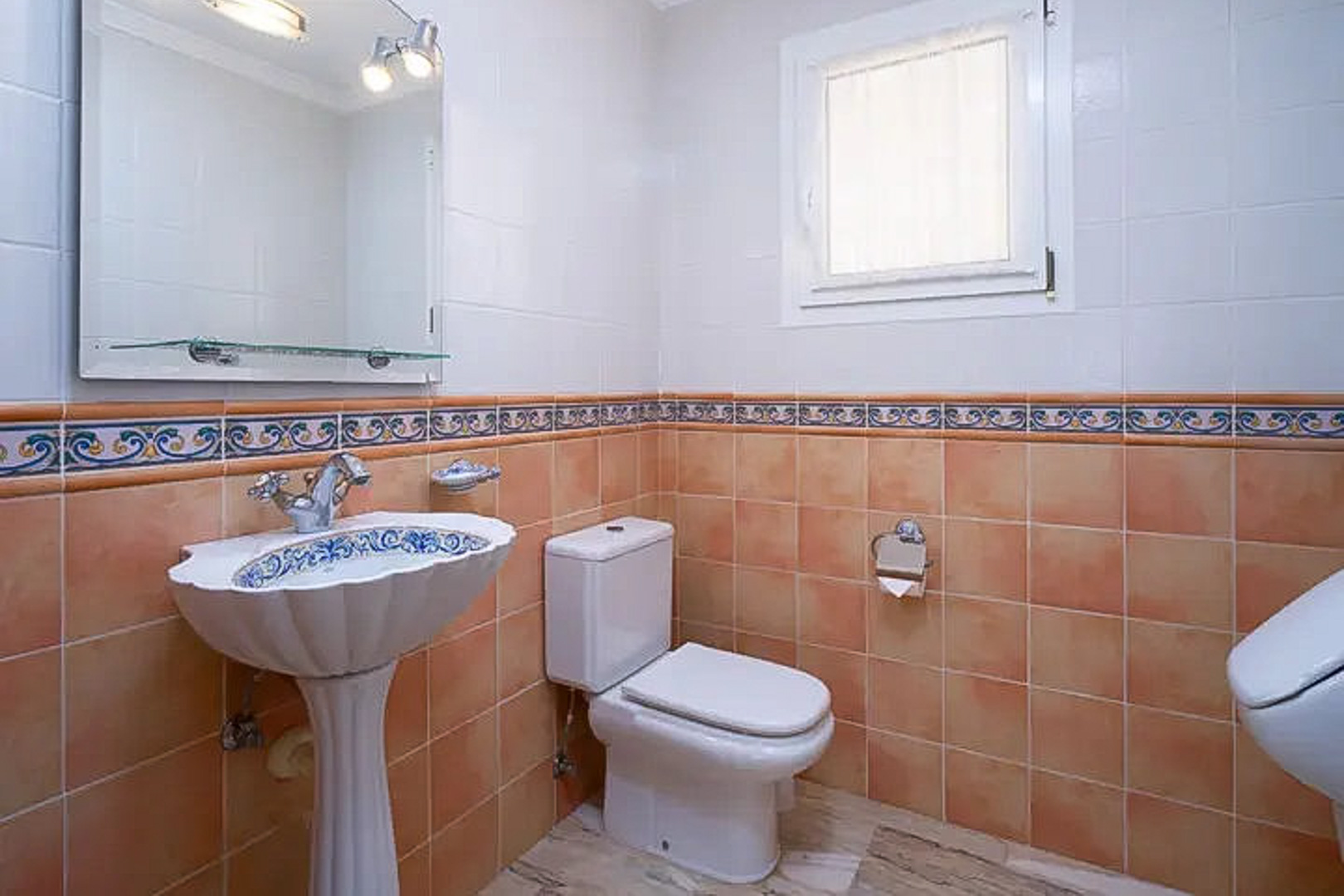 Guest bathroom featuring a decorative pedestal sink, terracotta and white wall tiling with an ornamental border, a large mirror, and a small frosted window.