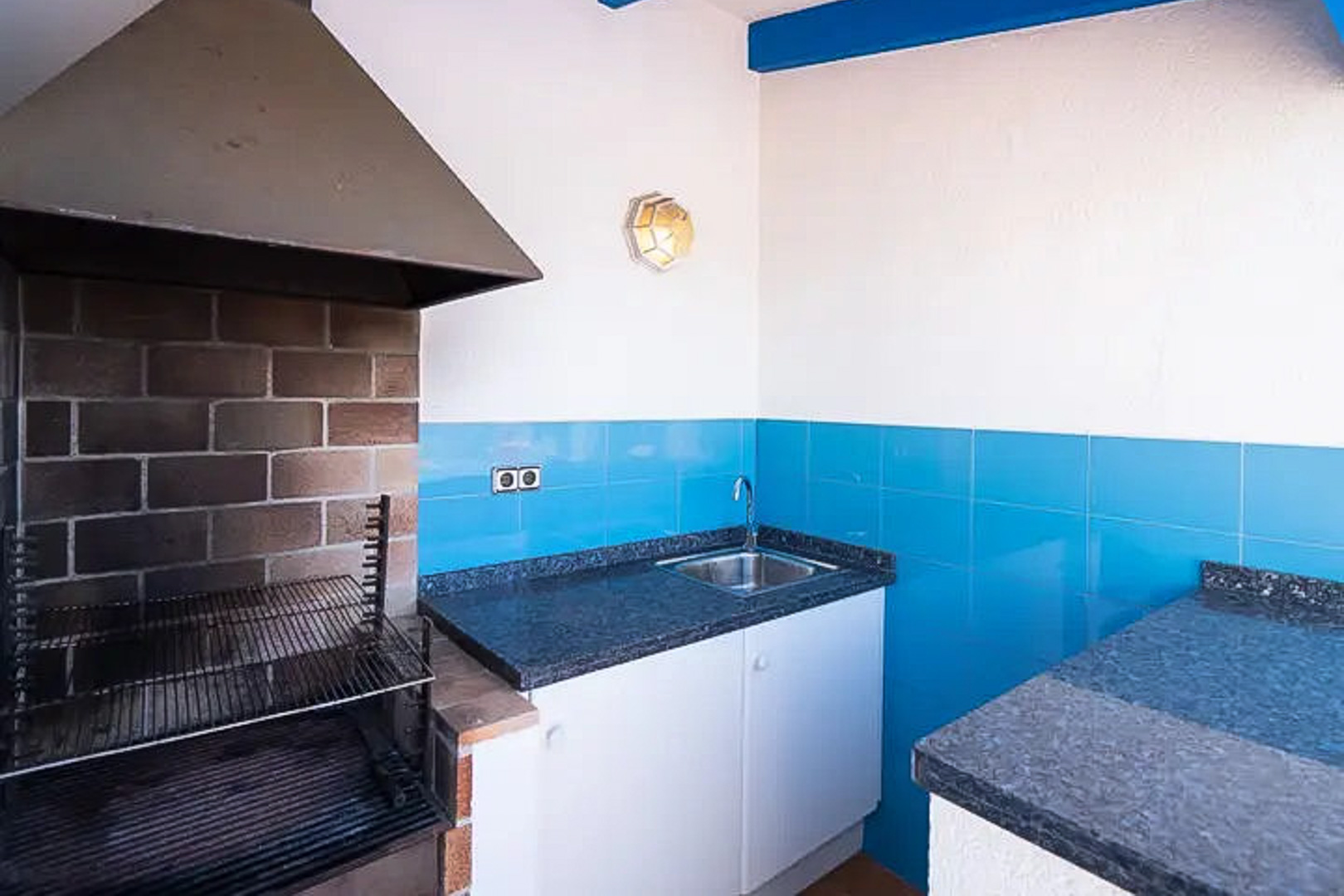 Outdoor barbecue area featuring a metal hood, dark granite countertops, a stainless steel sink, and vibrant blue Mediterranean tiling.