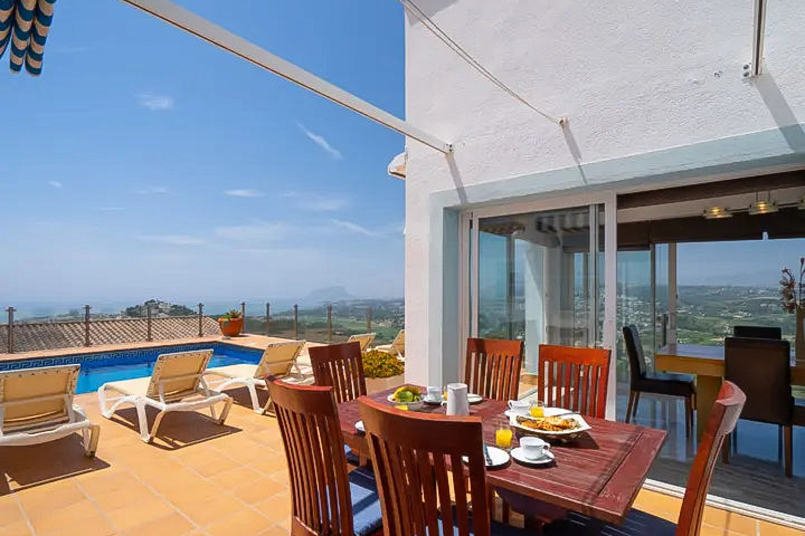 Outdoor terrace featuring a wooden dining set, private pool with decorative tiling, and panoramic views of the Mediterranean coastline and mountains.