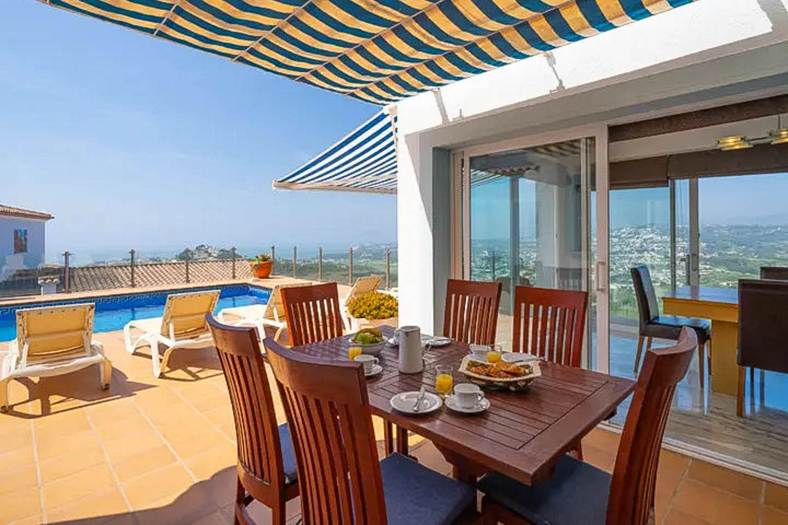 Outdoor terrace featuring a wooden dining set under a striped awning, overlooking the private pool and panoramic Mediterranean coastline.