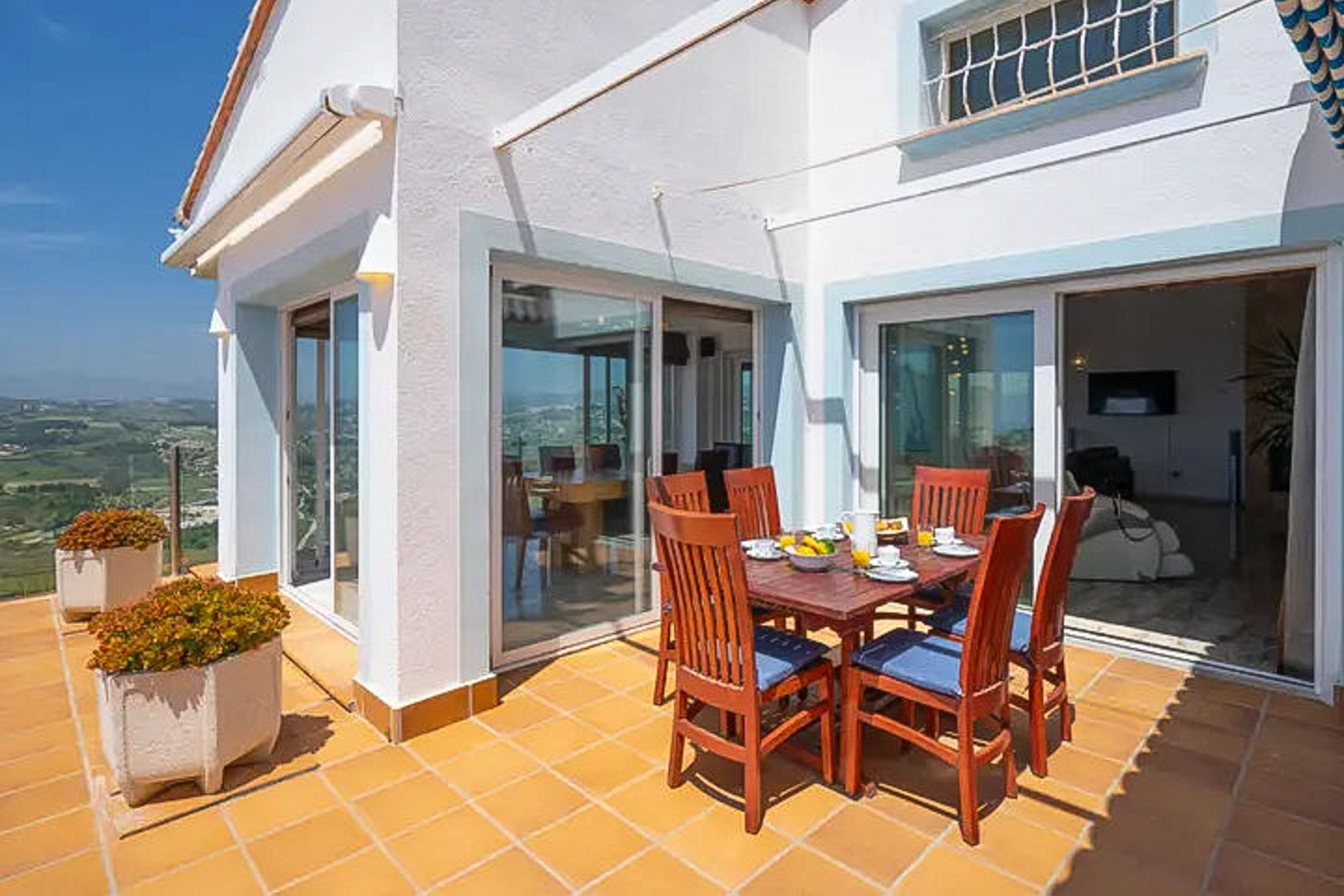 Outdoor terrace with terracotta flooring, a wooden dining set for six, and large sliding glass doors offering views of the surrounding green valley.