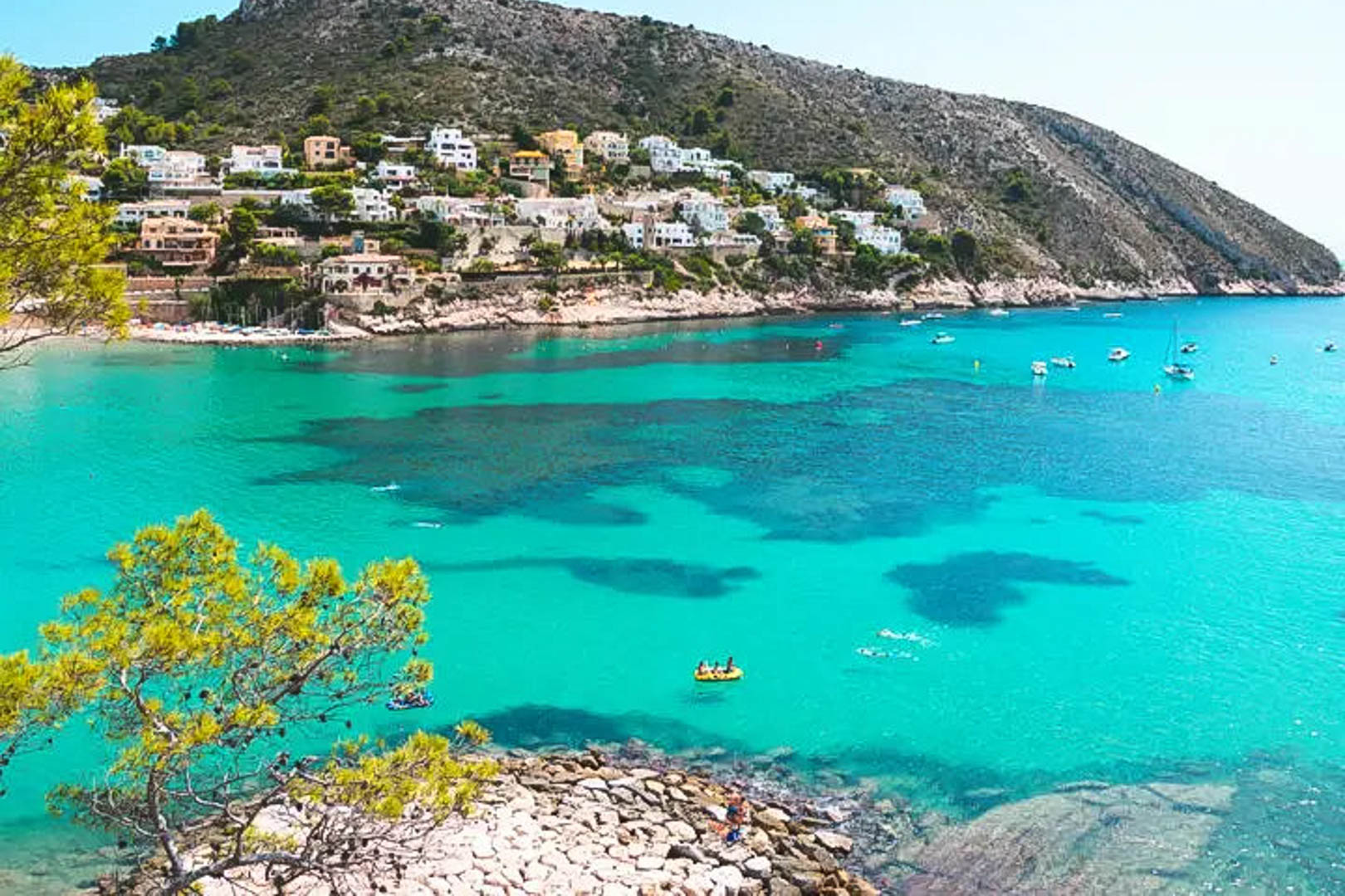 Panoramic view of a Moraira cove featuring turquoise waters, Mediterranean pines, and white villas nestled along the rocky coastline under a clear sky.