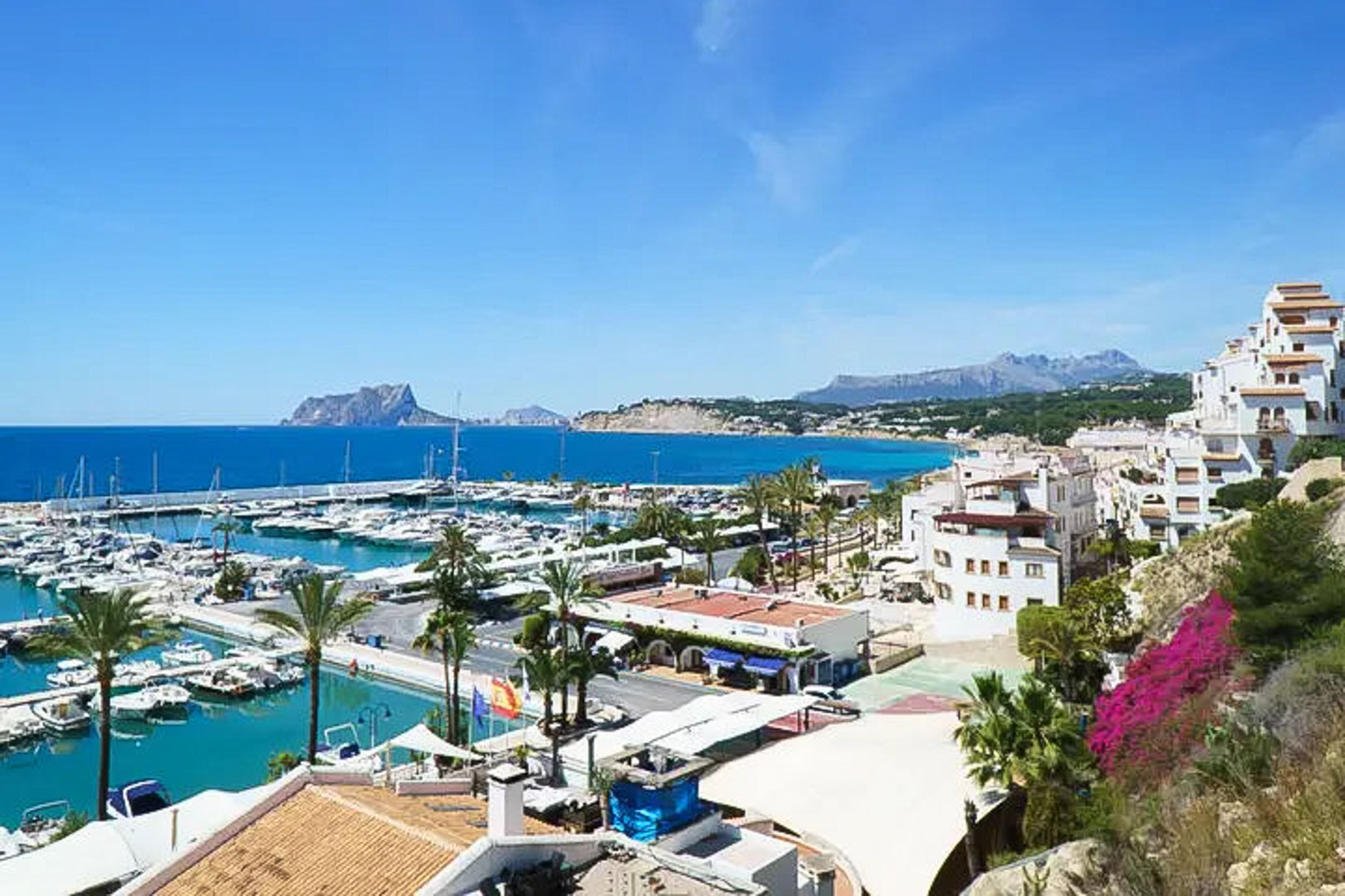 Panoramic view of Moraira marina featuring the Peñón de Ifach on the horizon, traditional white coastal architecture, and turquoise Mediterranean waters.