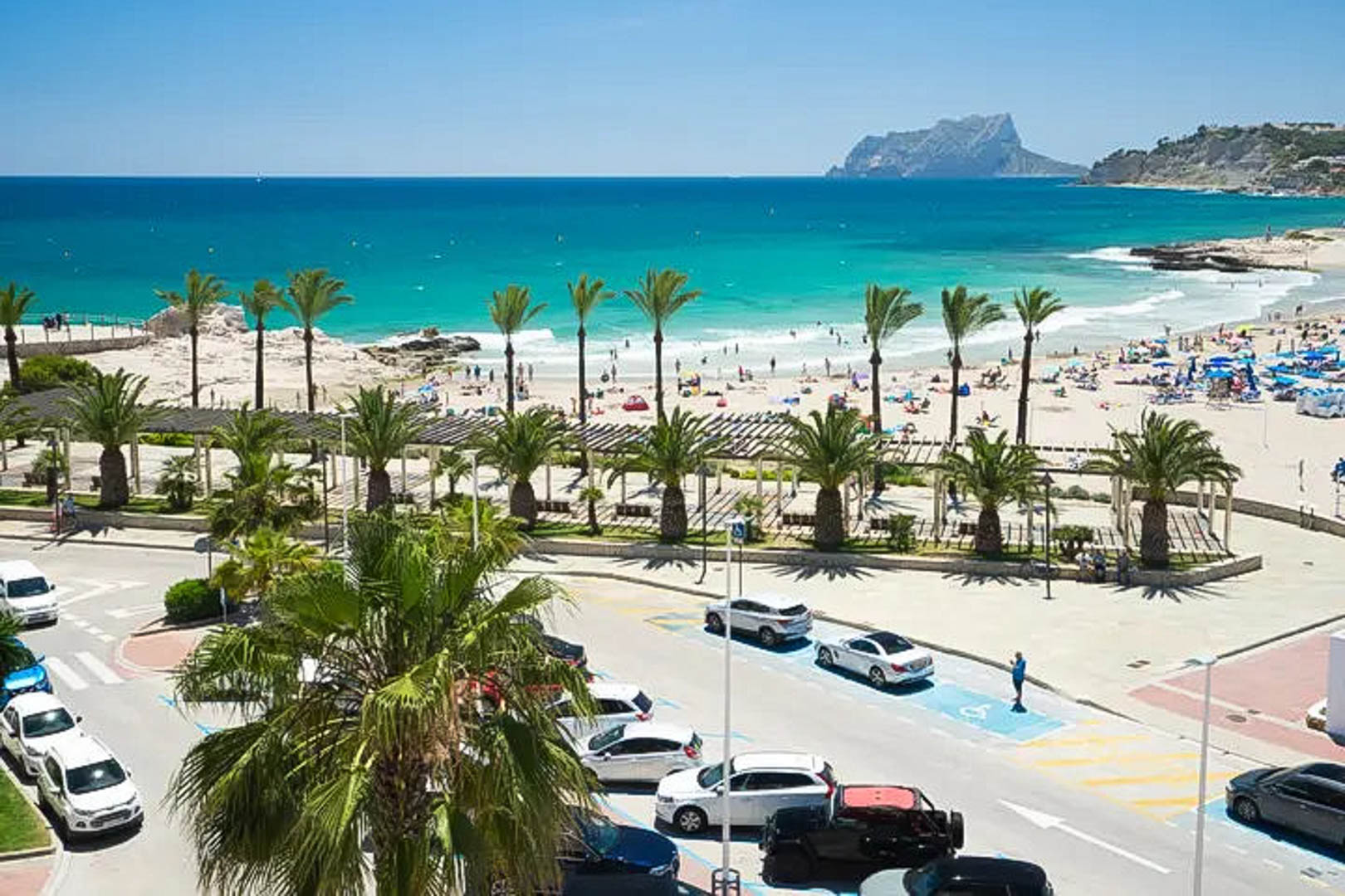 Panoramic view of Moraira's sandy beach and turquoise Mediterranean waters, featuring the Peñón de Ifach landmark and a palm-lined coastal promenade.