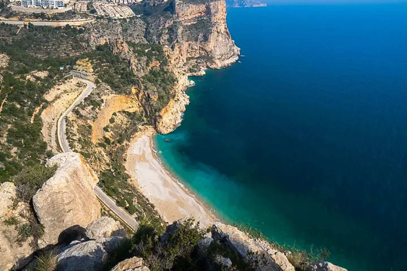Panoramic view of the Costa Blanca cliffs, featuring a turquoise cove and a winding coastal road overlooking the Mediterranean Sea.