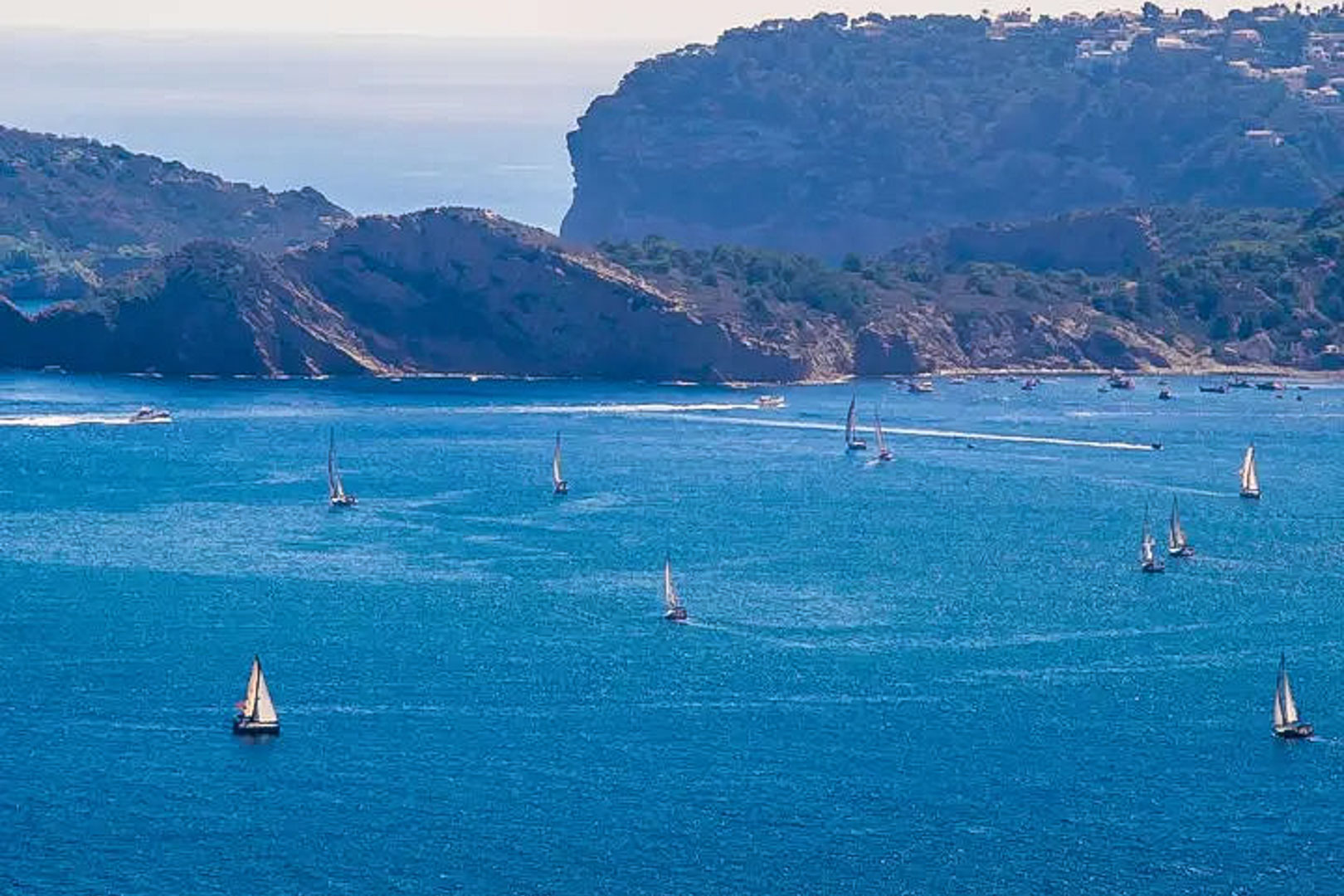 Panoramic view of the Mediterranean Sea in Moraira, featuring several sailboats navigating near the rocky cliffs of the Costa Blanca coastline.