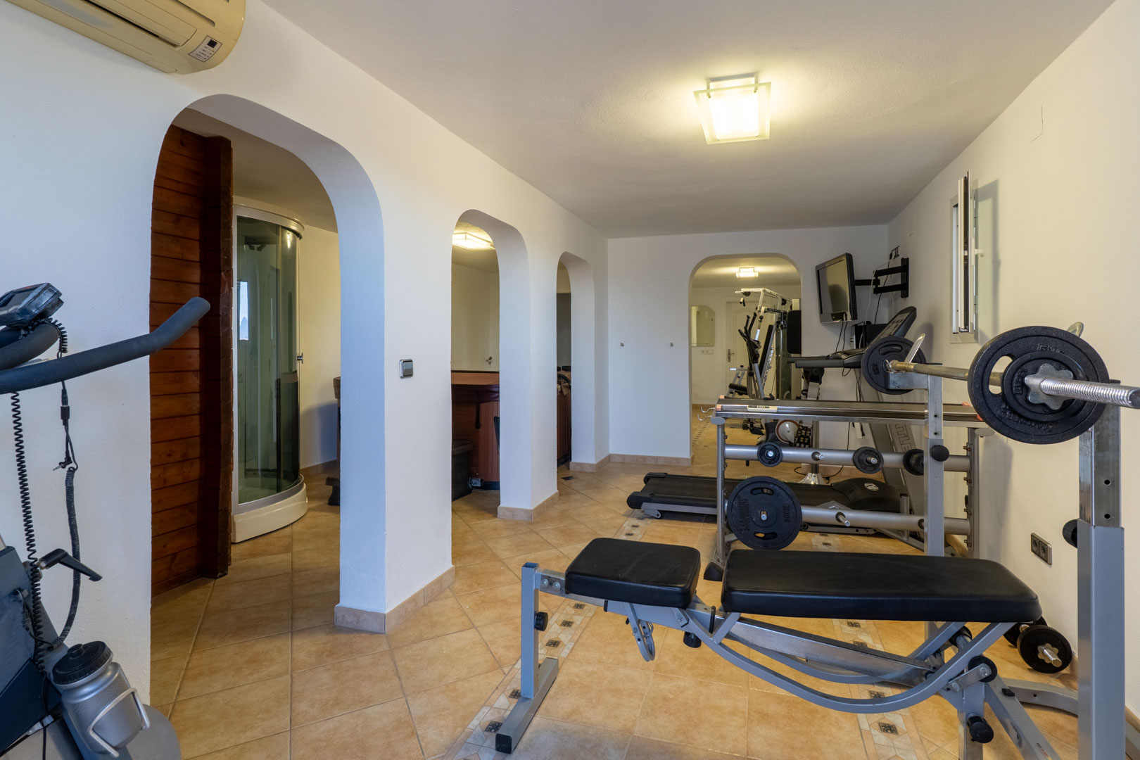 Private home gym and wellness suite featuring weight training equipment, a treadmill, and arched entrances leading to the sauna, steam shower, and hot tub.