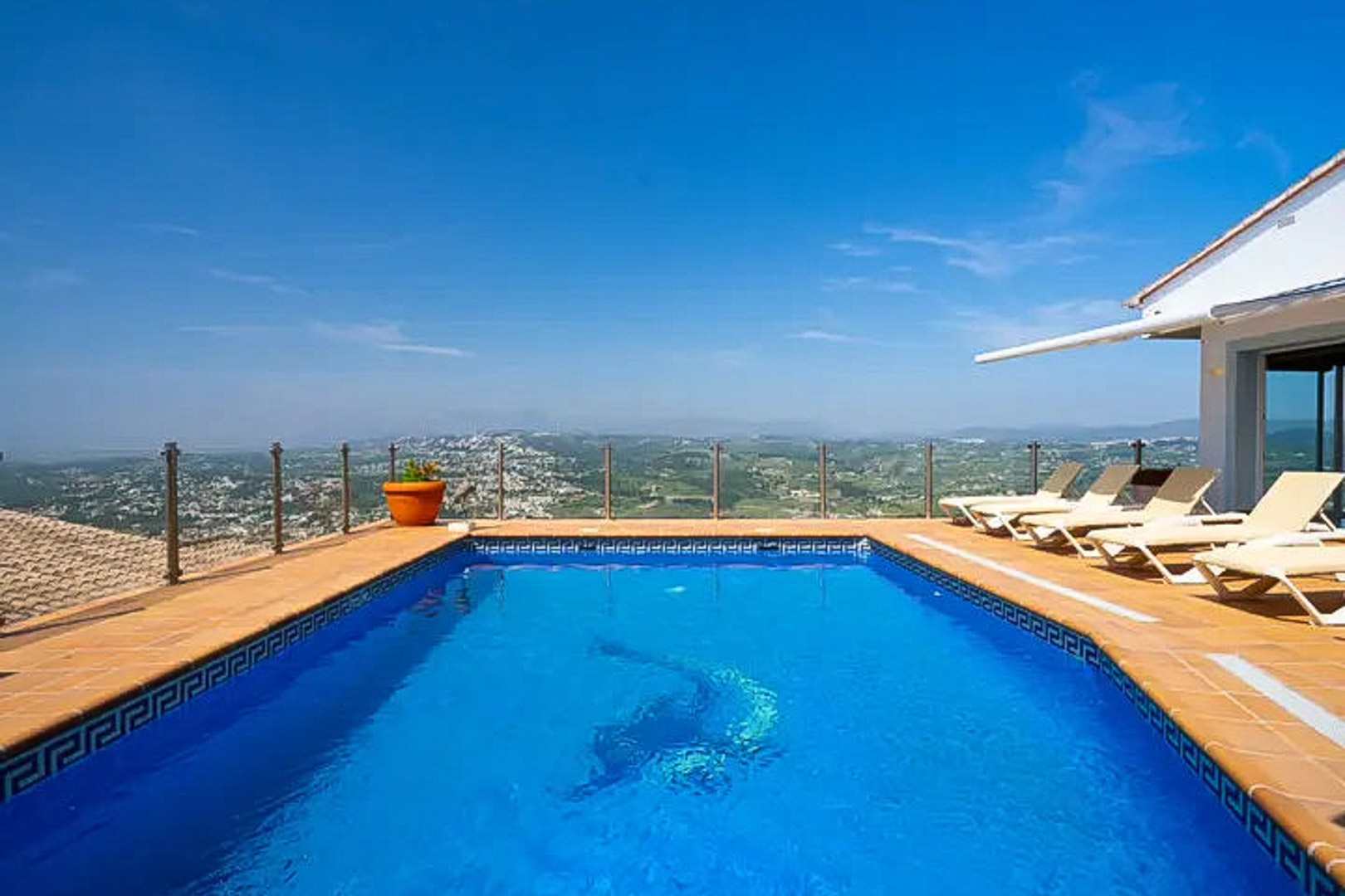 Rectangular pool featuring a dolphin mosaic, sun deck with loungers, and a glass railing overlooking the expansive coastal and mountain landscape.