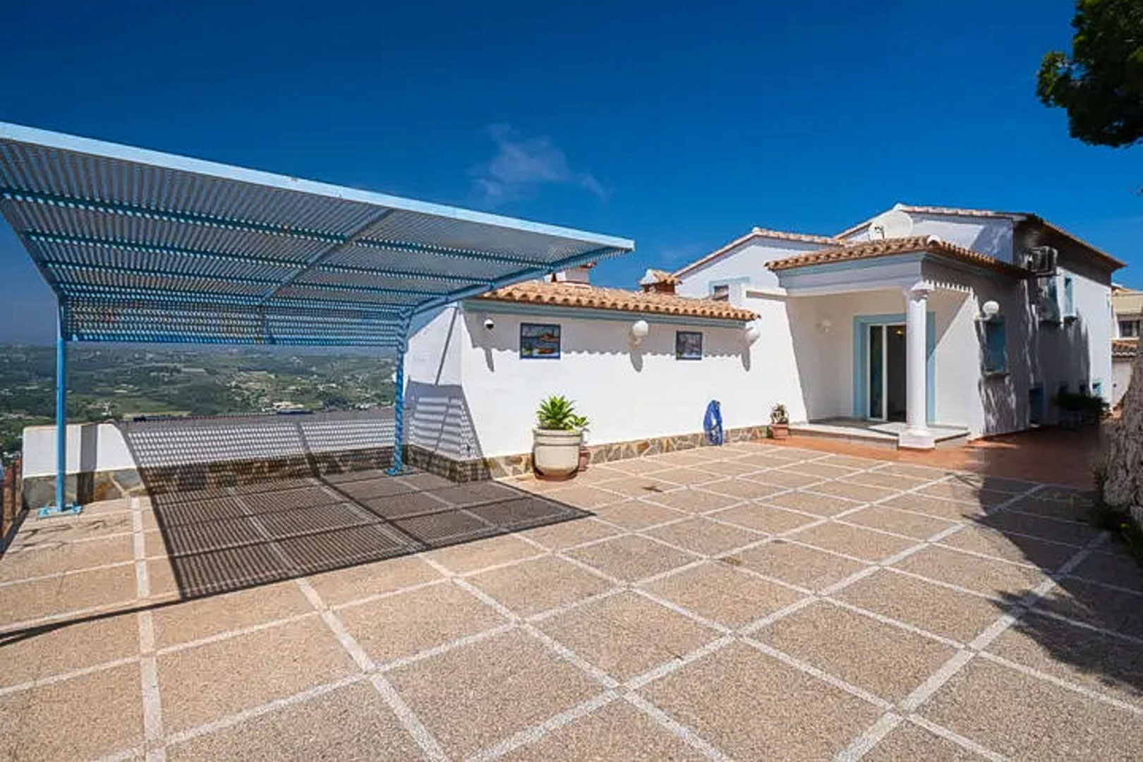 Spacious outdoor parking area featuring tiled flooring, a blue metal carport, and panoramic views of the surrounding Mediterranean hills and landscape.