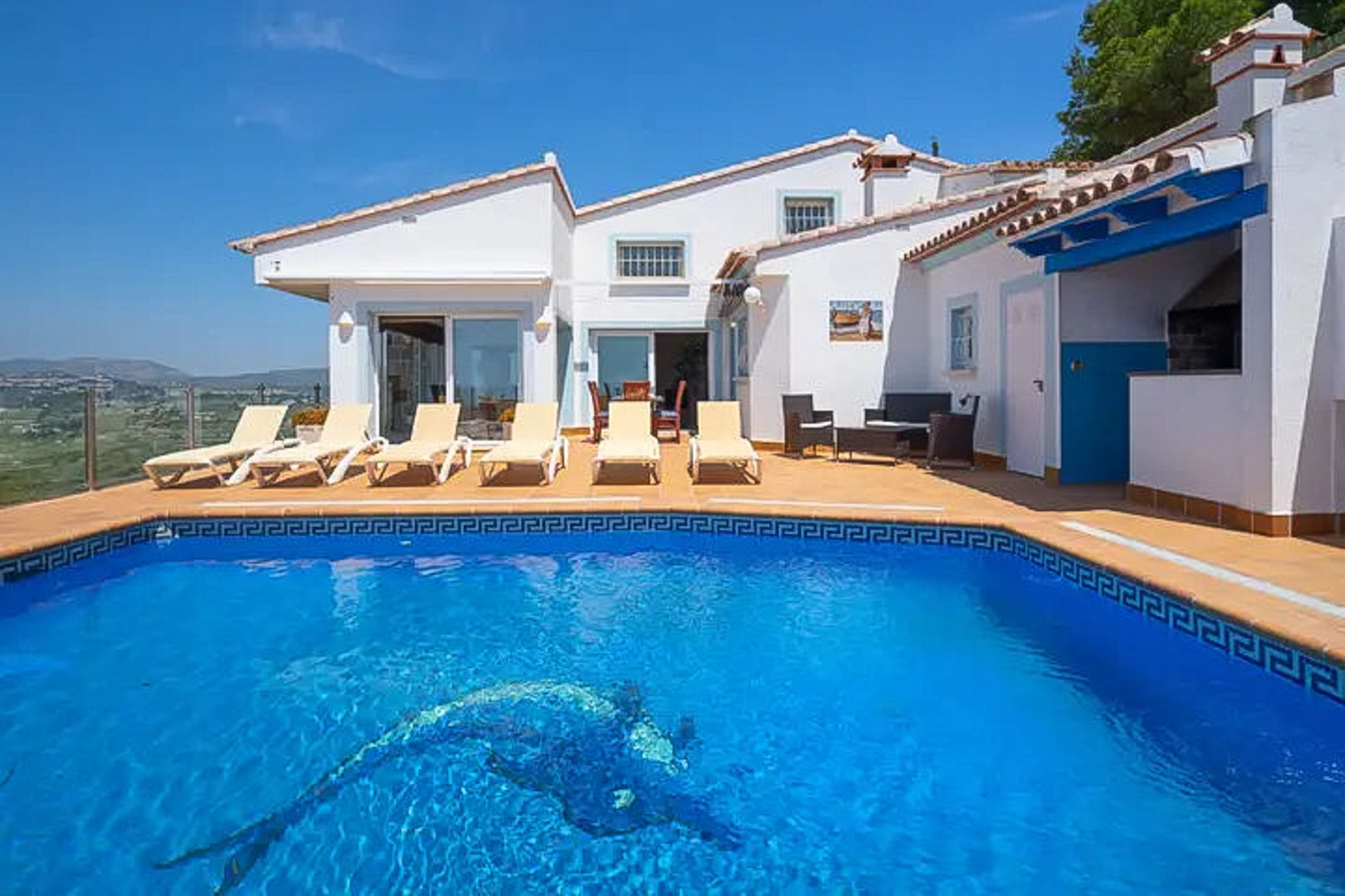 Terrace featuring a pool with a dolphin mosaic, sun loungers, and a built-in barbecue area under blue wooden beams, overlooking the valley.
