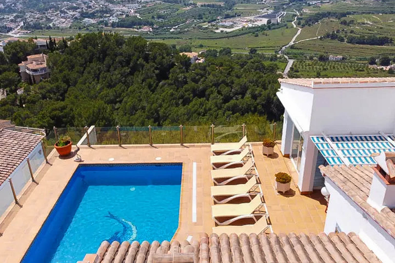 Terrace featuring a rectangular pool with dolphin mosaic, sun loungers, and glass railings overlooking the lush valley and mountains of Moraira.