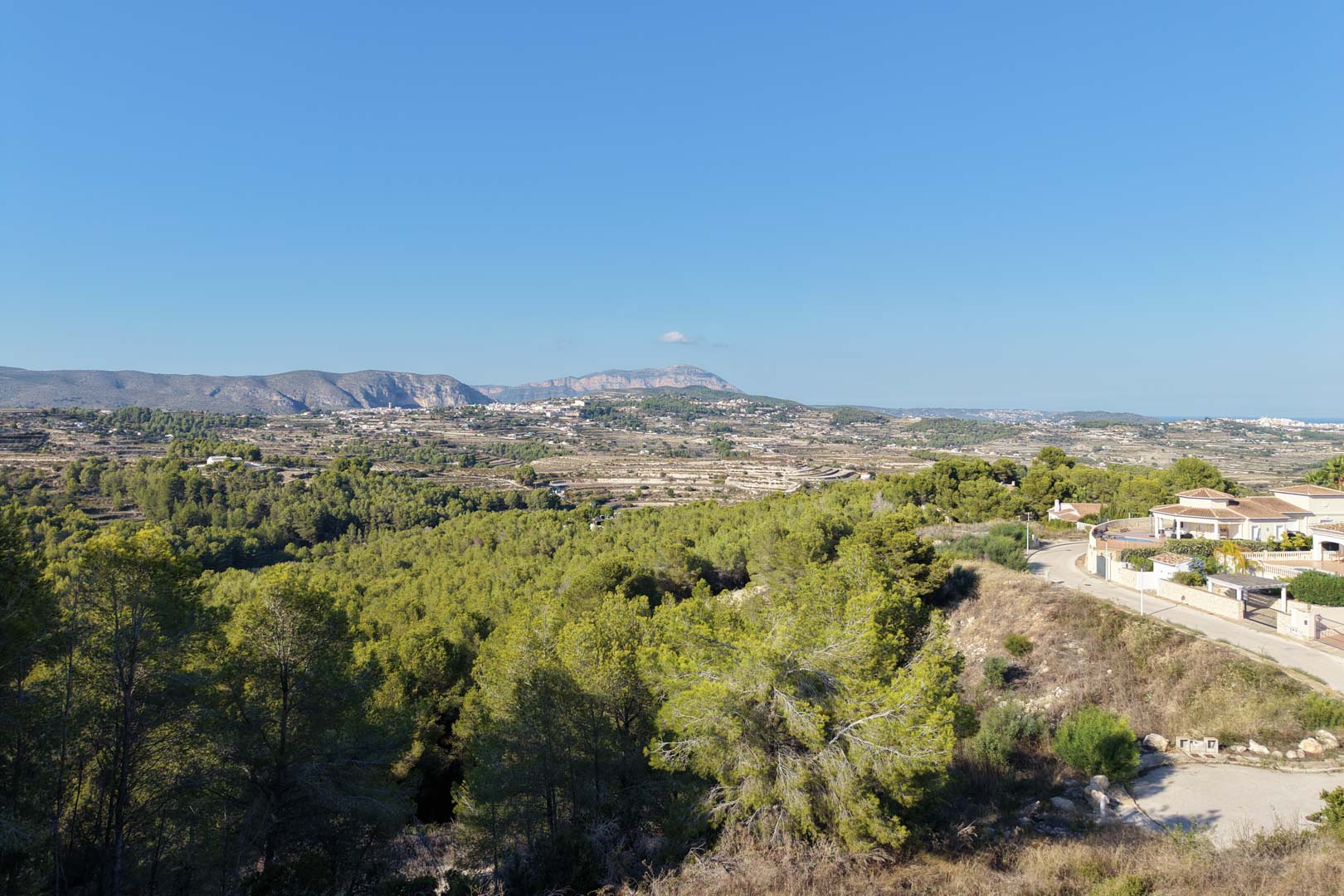 Panoramic open views overlooking the valley and surrounding mountains, bordered by dense Mediterranean pine forests under a clear blue sky.