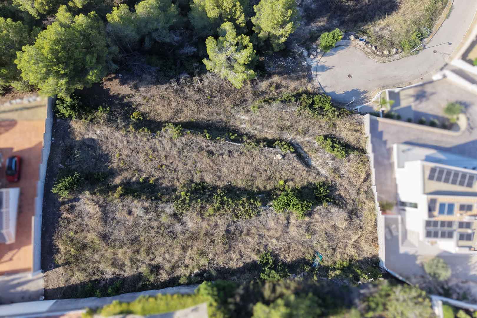 Zenithal aerial view of the terraced urban plot featuring dry stone walls and Mediterranean scrub, located adjacent to modern villas and a paved cul-de-sac.