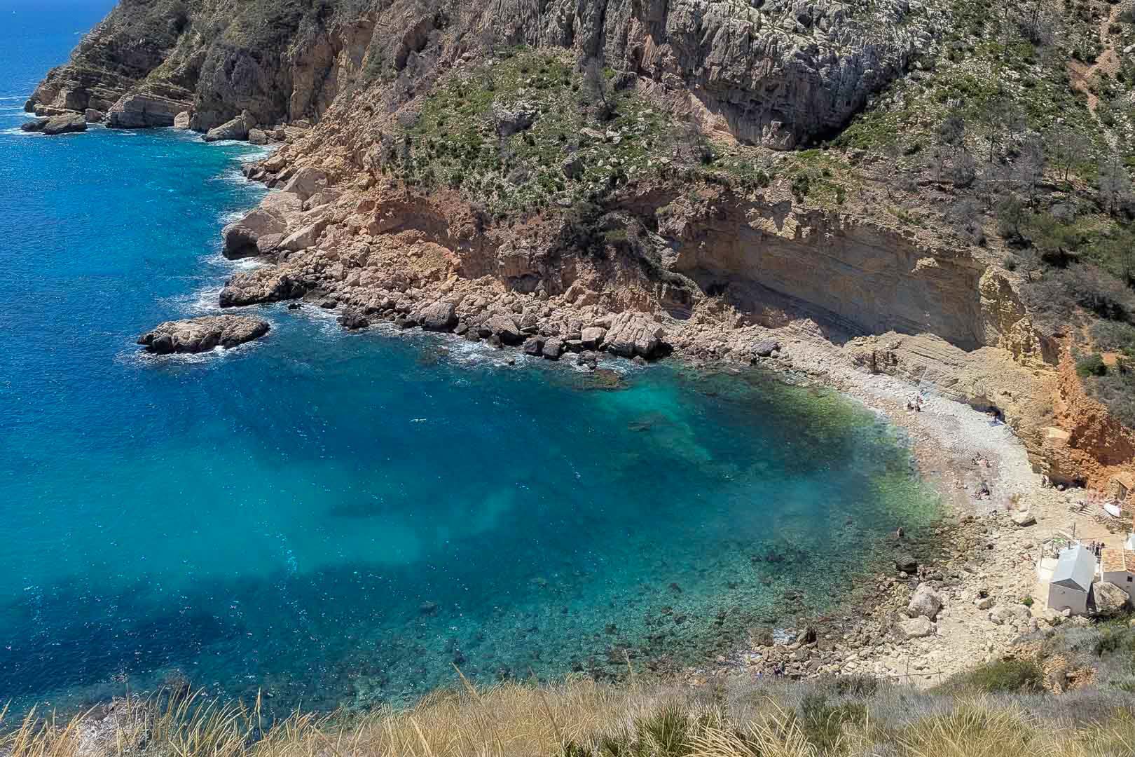 Aerial view of a Mediterranean cove featuring turquoise waters, rugged limestone cliffs, and a small pebble beach along the Moraira coastline.
