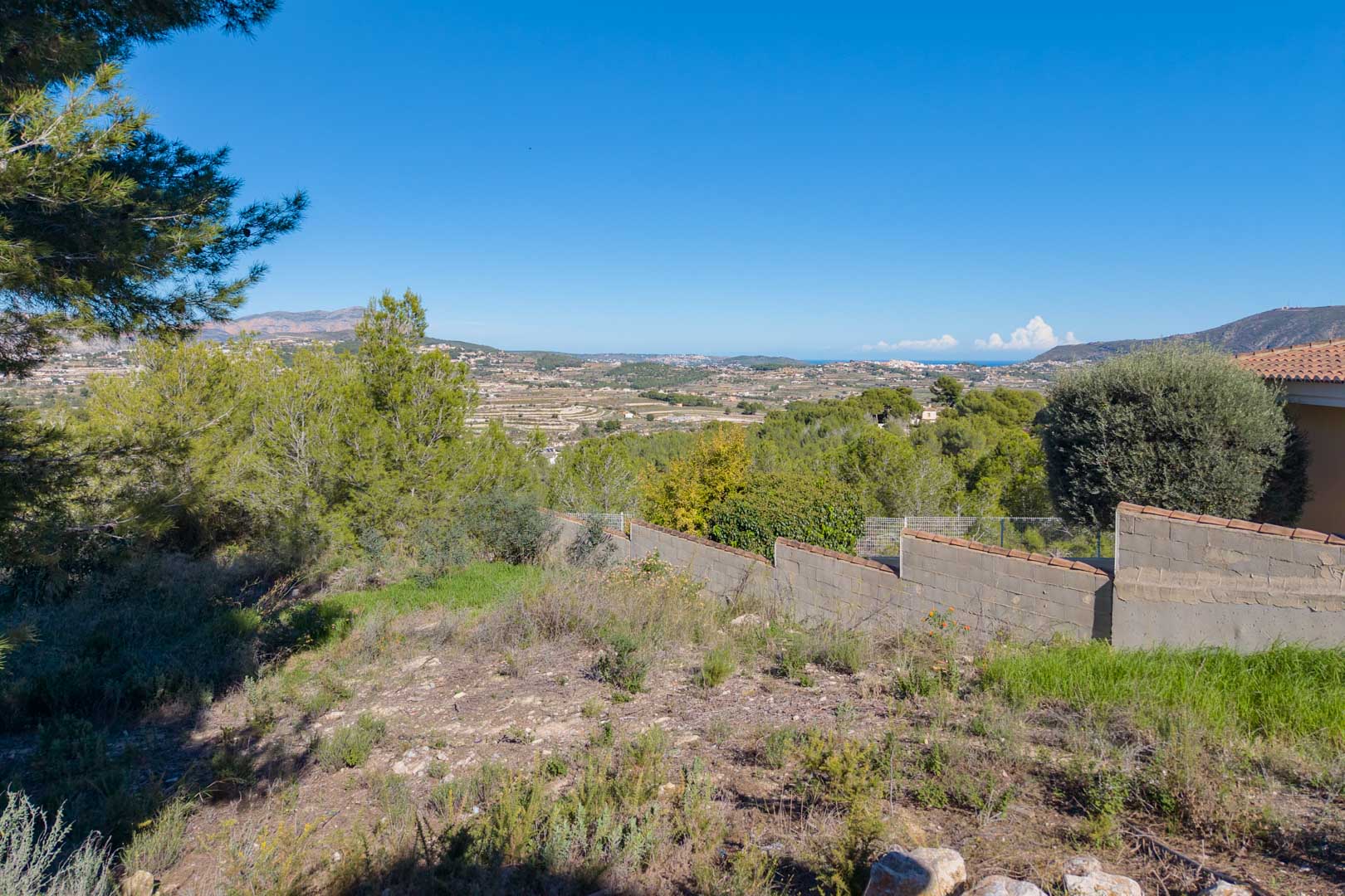 Elevated plot featuring panoramic valley and partial sea views. The terrain includes natural vegetation and a concrete boundary wall with tile capping.