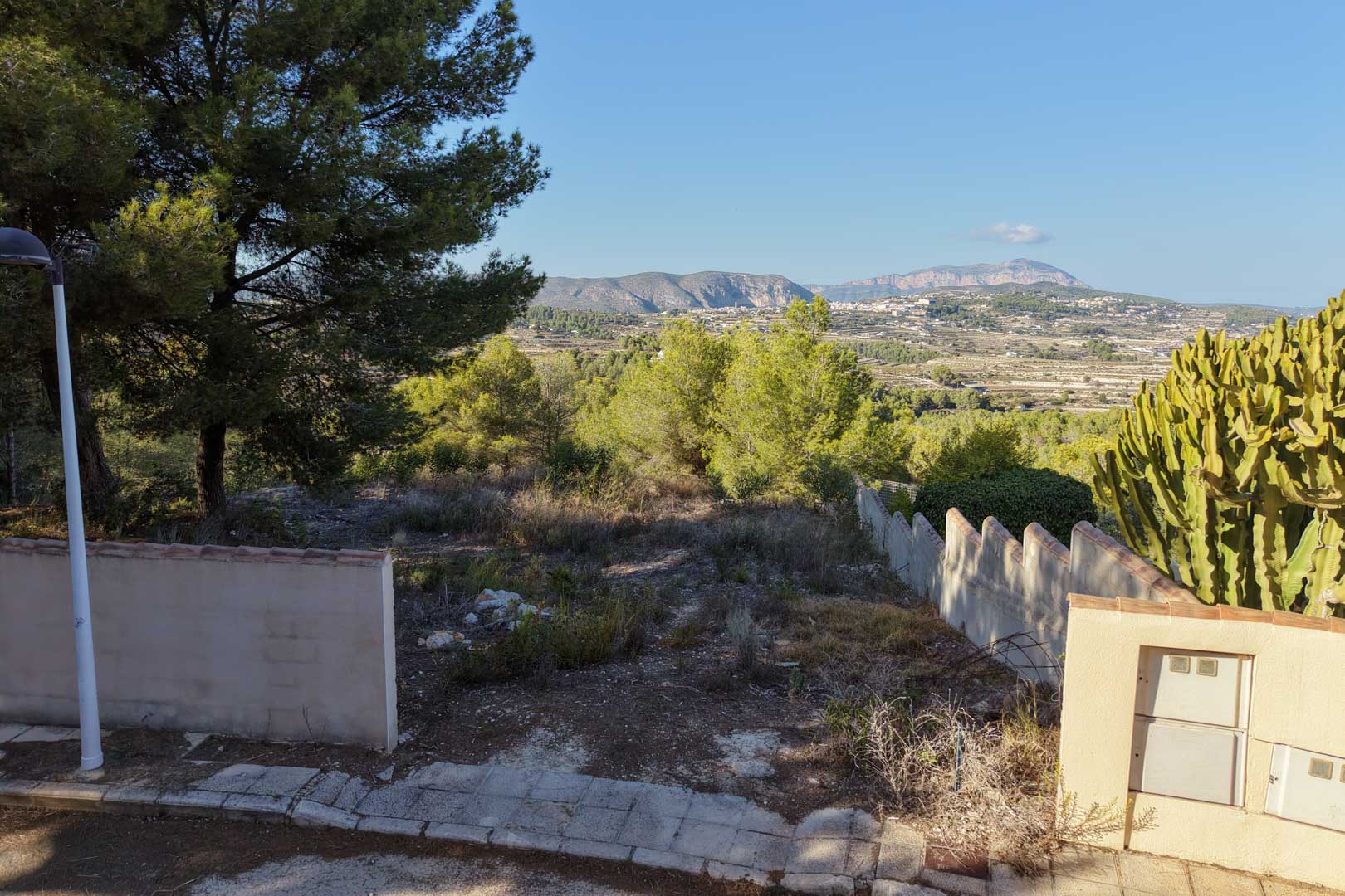 Elevated urban plot in Benimeit featuring panoramic valley and mountain views. The site includes perimeter walls, native vegetation, and direct access from a paved residential street.