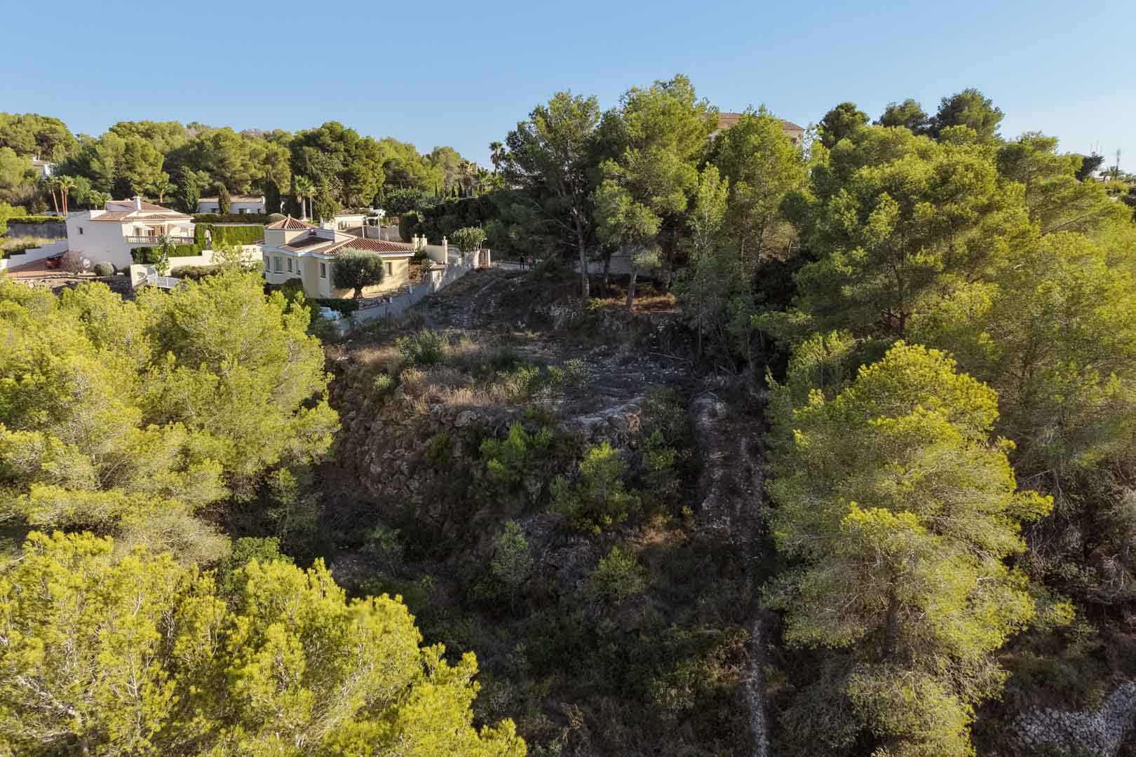 Elevated urban plot in Benimeit, surrounded by mature pine trees and Mediterranean vegetation, featuring natural terraced terrain and views of neighboring villas.