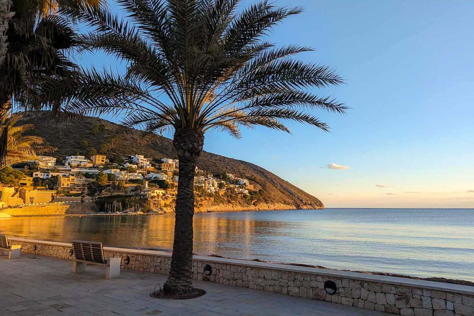 Moraira's coastal promenade featuring stone benches and palm trees, overlooking the calm bay and hillside residences during the golden hour.