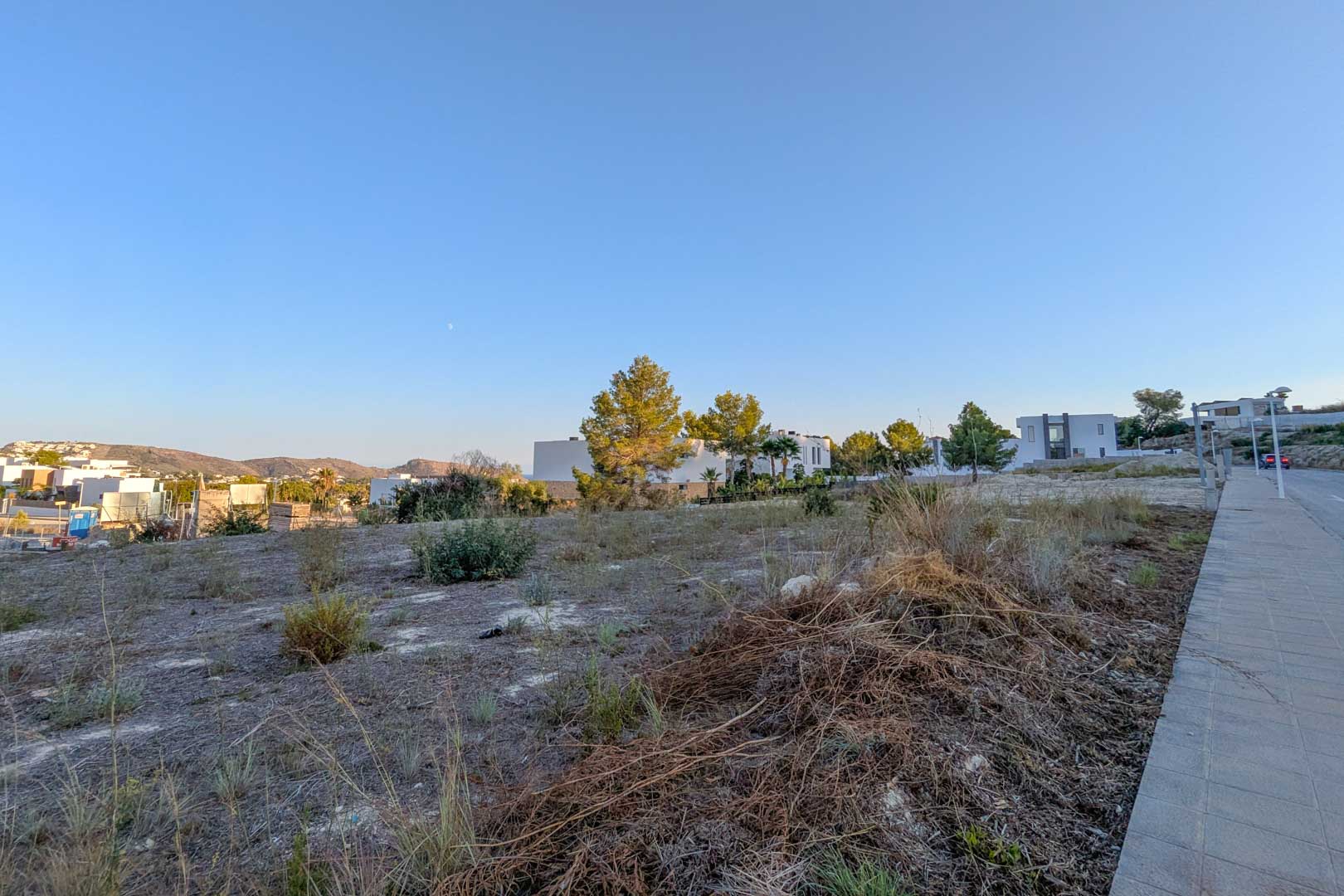 Flat urban plot in El Tesoro, Moraira, featuring open valley views and proximity to modern villas. The land includes direct access via a paved sidewalk and street.