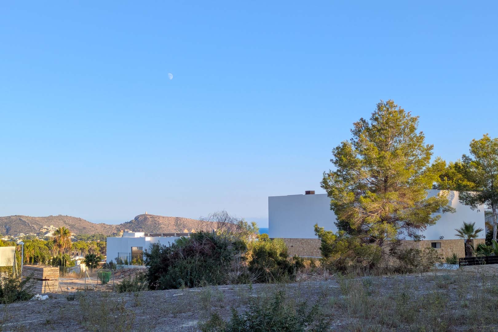 View from the plot overlooking modern cubic villas and Mediterranean pines, with distant mountain ridges and a glimpse of the sea.