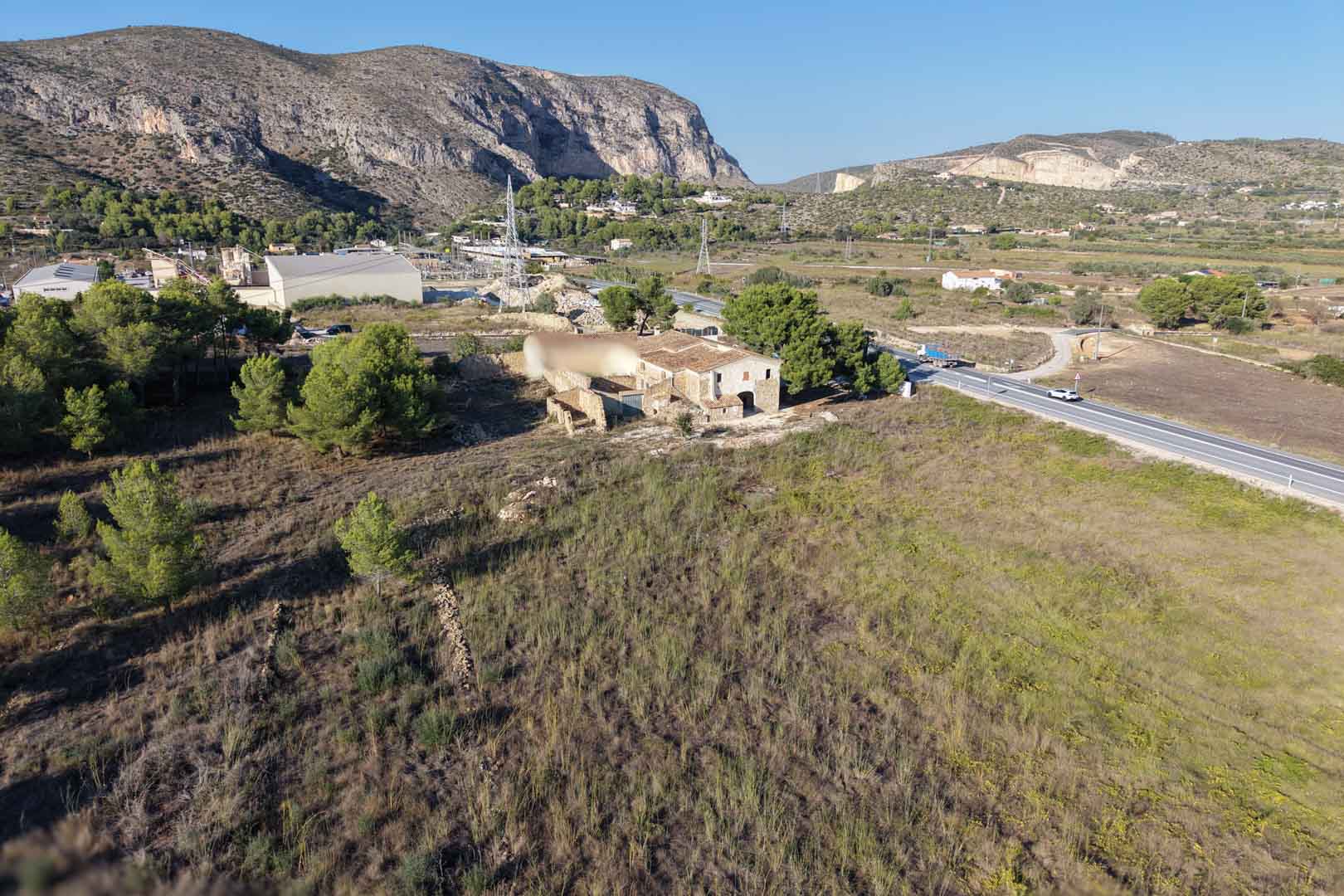 Aerial perspective of the traditional stone farmhouse on a vast flat plot in Teulada, featuring mountain backdrops and easy road access.