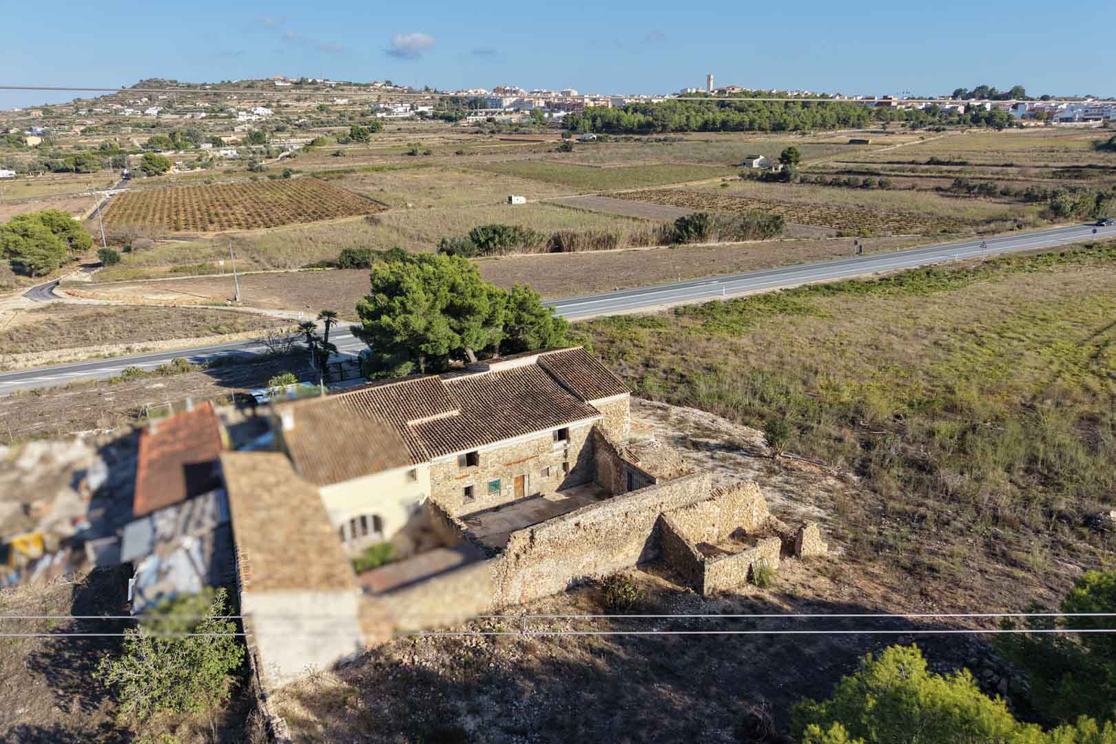 Aerial view of the traditional stone farmhouse and its outbuildings, set on an expansive rural plot with clear views of the Teulada skyline.