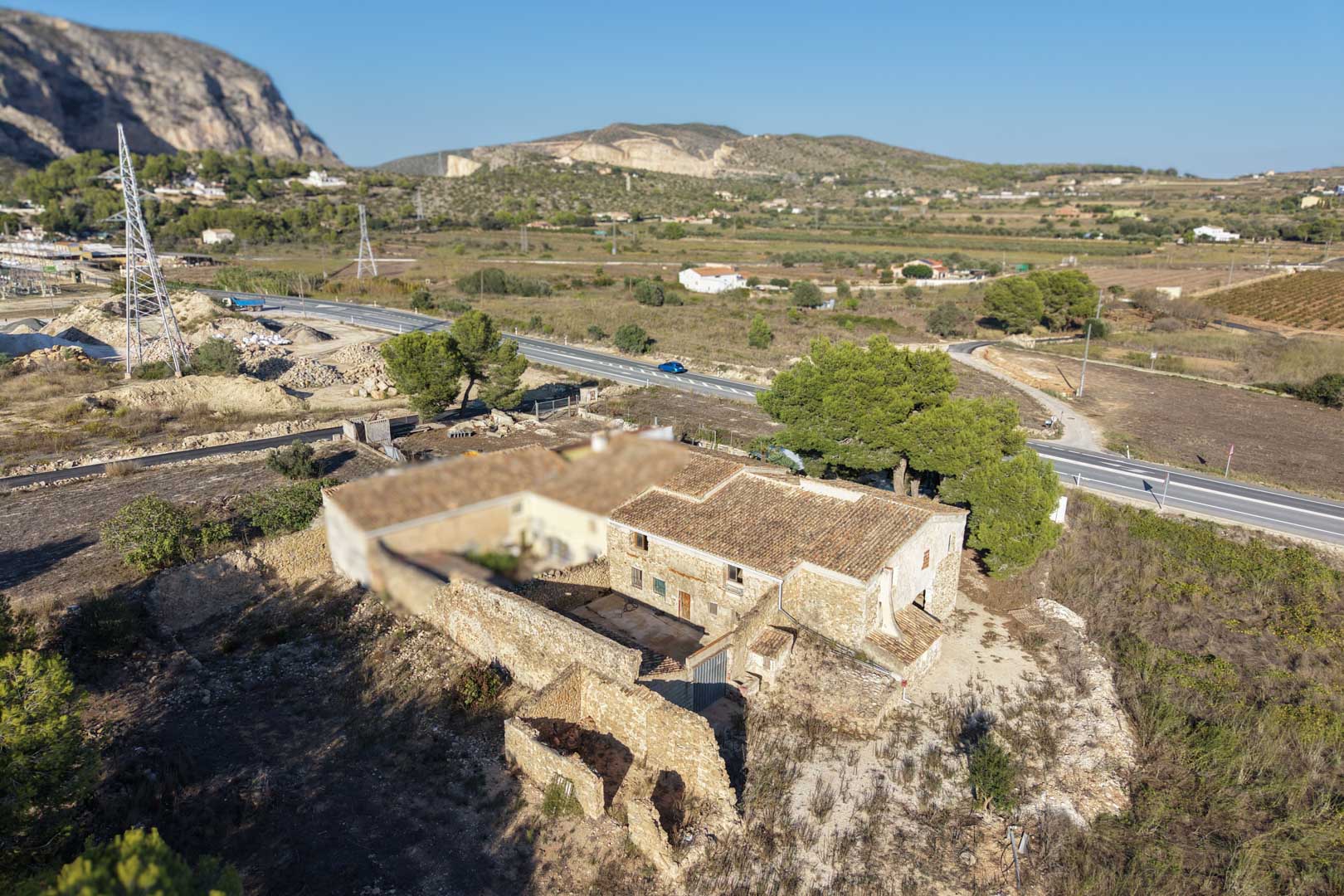 Aerial view of the traditional stone farmhouse featuring a terracotta tile roof, a central courtyard, and a flat plot set against a mountainous rural landscape.