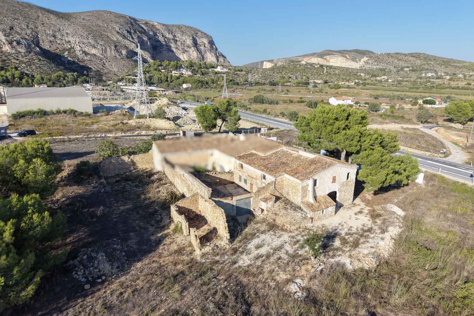 Aerial view of the traditional stone farmhouse featuring an inner courtyard and outbuildings, set on a vast rustic plot with views of the Teulada mountains.