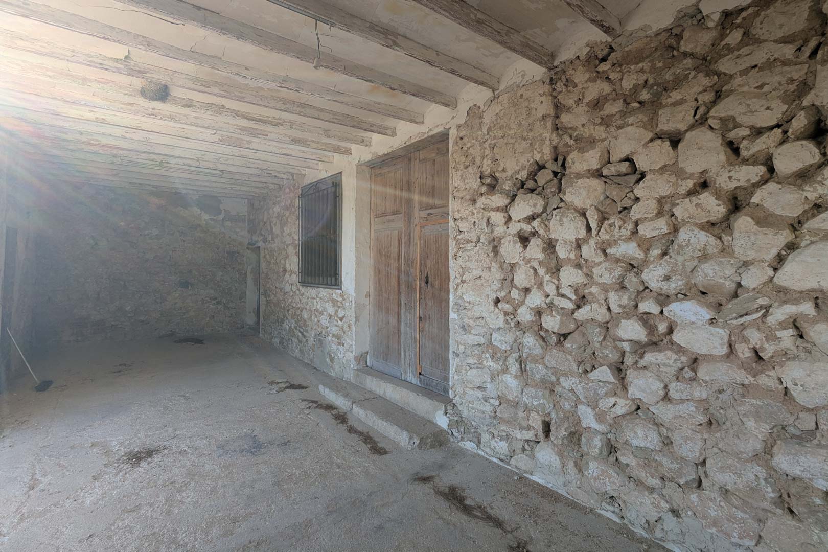 Covered porch featuring exposed natural stone walls, original wooden ceiling beams, and a traditional double wooden entrance door characteristic of the region.
