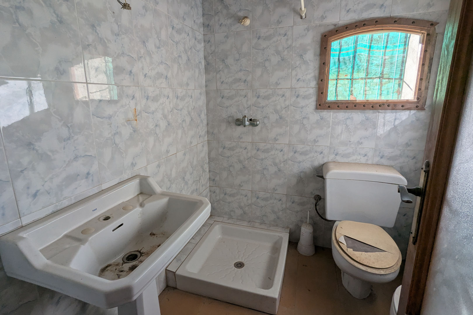 Original bathroom featuring blue marble-effect wall tiles, a pedestal sink, square shower tray, and a small arched window with a wooden frame.