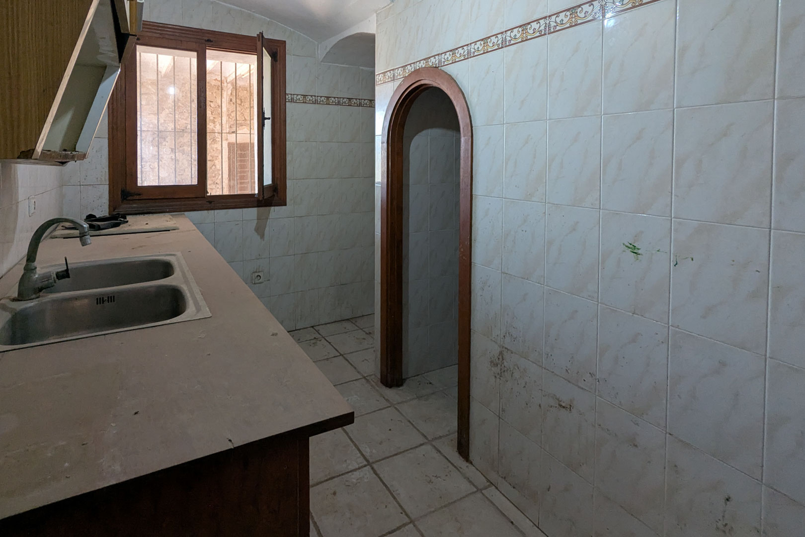 Original rustic kitchen featuring a double stainless steel sink, tiled walls with decorative borders, and a narrow arched wooden doorway.
