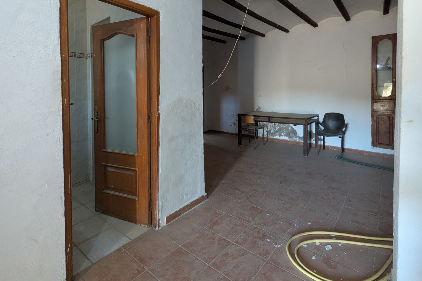 Rustic interior featuring exposed dark wood ceiling beams, terracotta floor tiles, and lime-washed walls. A wooden door with glass and a built-in cupboard are visible.
