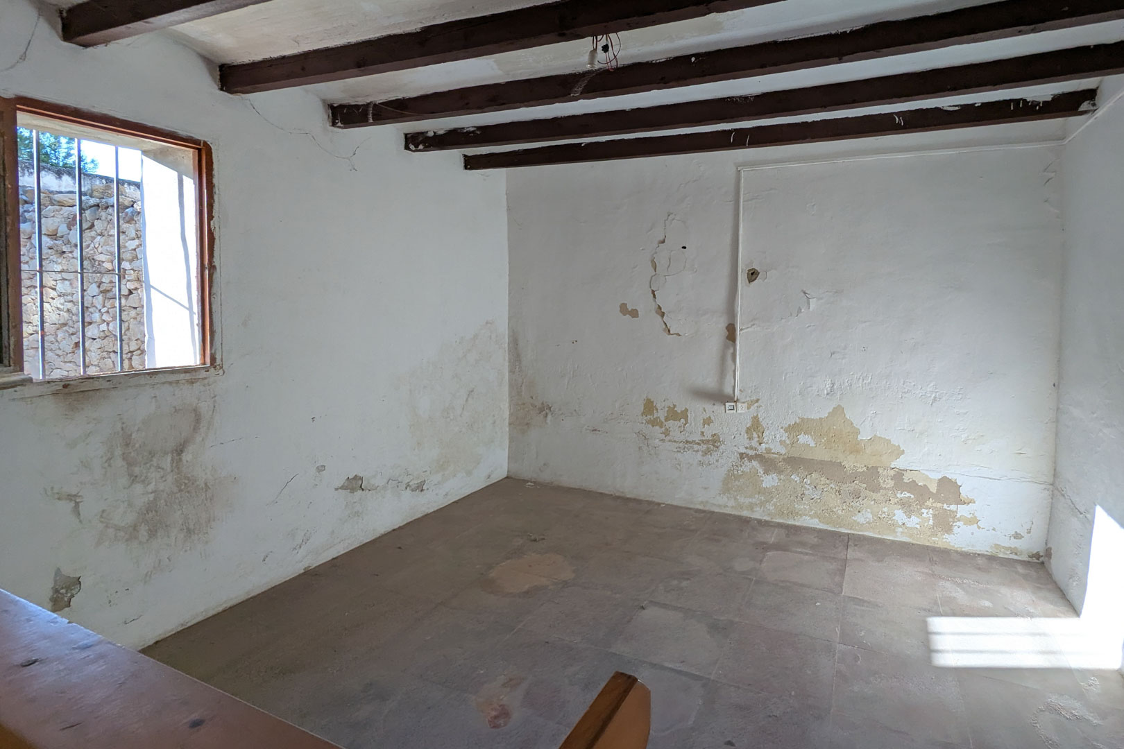 Rustic interior featuring original dark wood ceiling beams, lime-washed walls with authentic wear, and traditional tiled flooring. A barred window provides natural light and stone wall views.