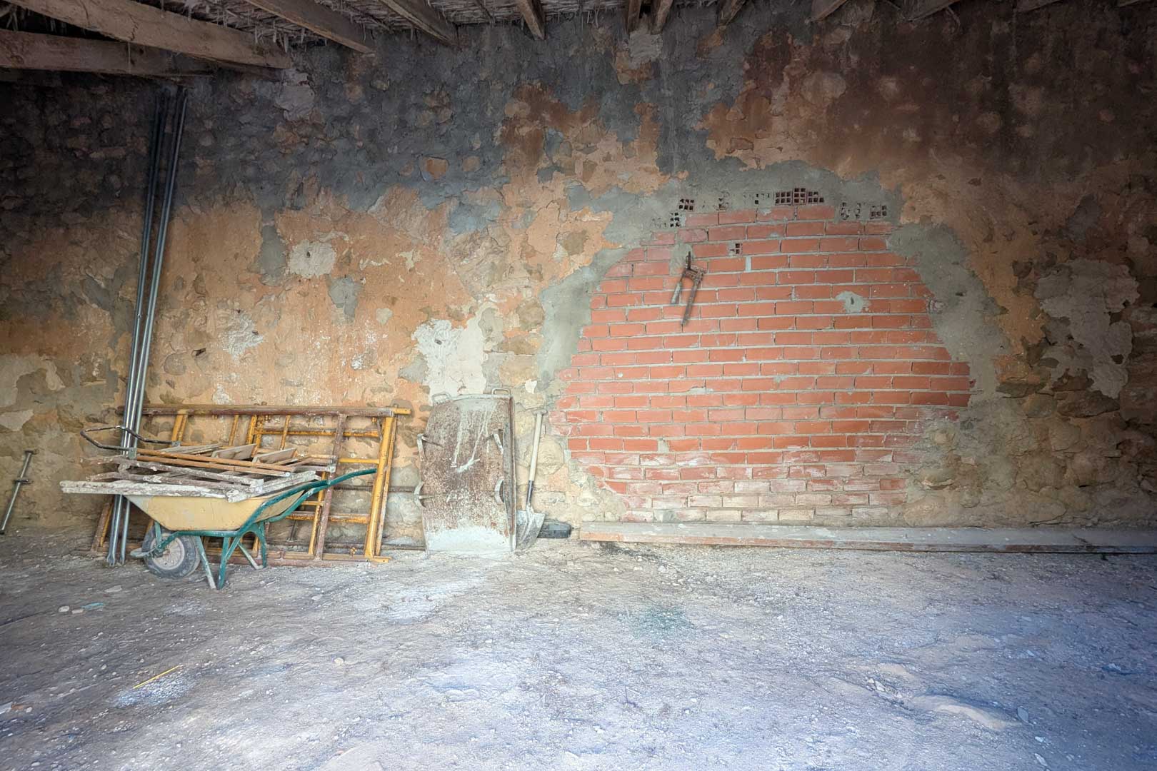 Rustic interior featuring original stone walls with a brick repair, exposed wooden beams, and a traditional cane ceiling. The space currently houses construction tools.