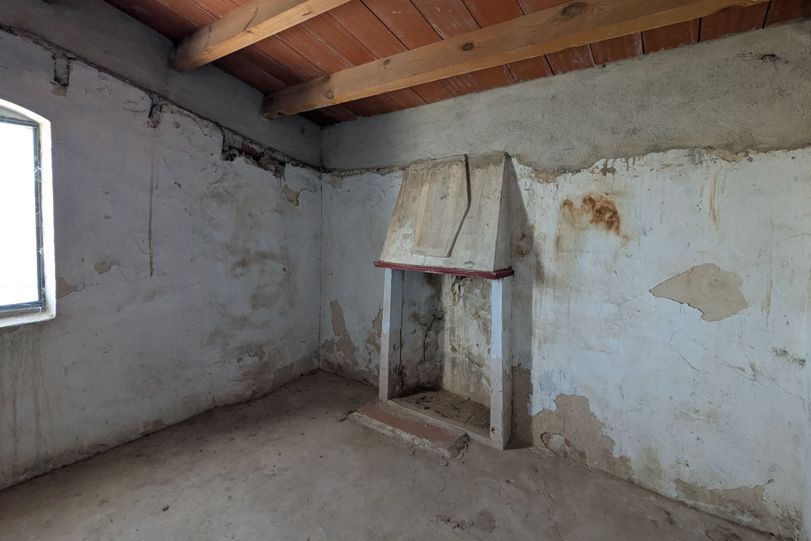 Rustic living area featuring a traditional hooded fireplace, exposed wooden ceiling beams, and original lime-washed walls awaiting restoration.