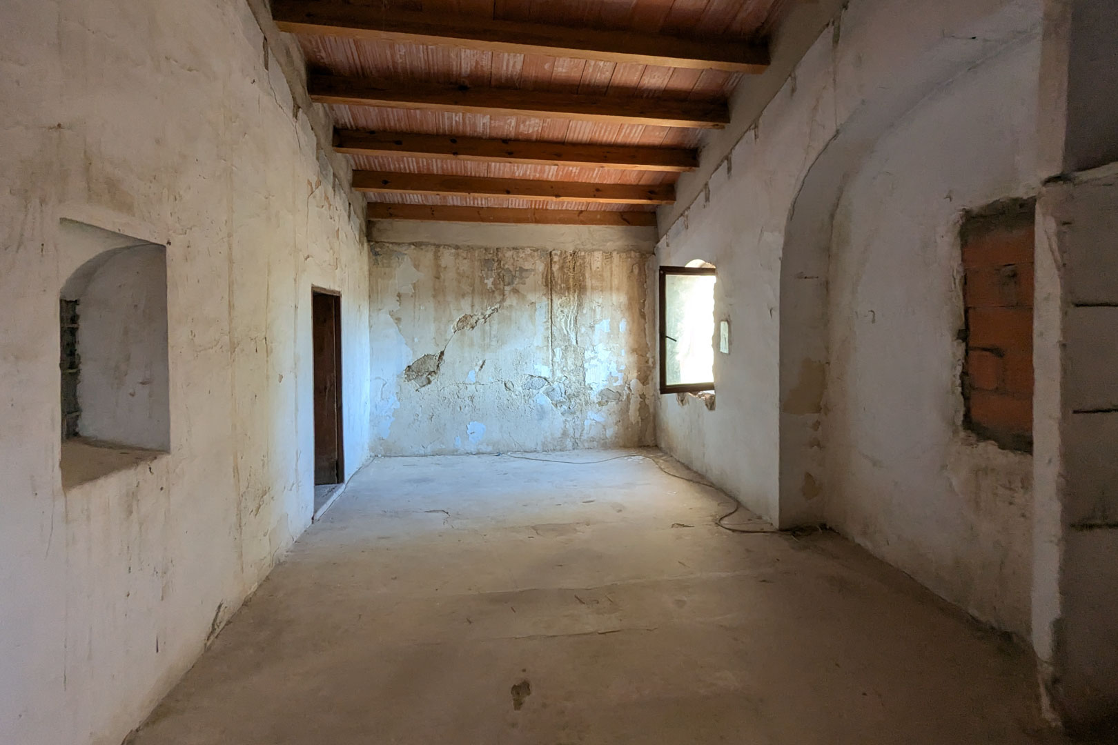 Spacious open-plan room featuring high ceilings with exposed wooden beams and terracotta tiles. Thick masonry walls include original arched niches and a window for natural light.