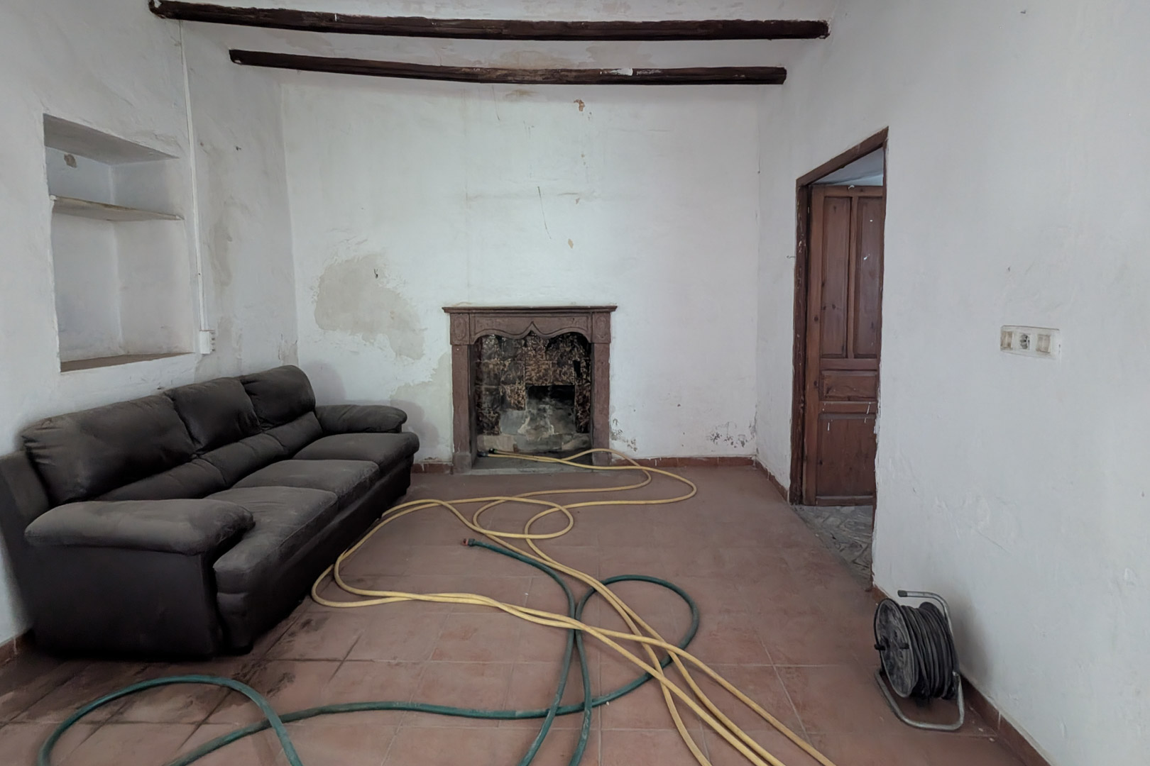 Traditional living room featuring exposed dark wood ceiling beams, terracotta tile flooring, and an original fireplace with a carved stone mantelpiece.
