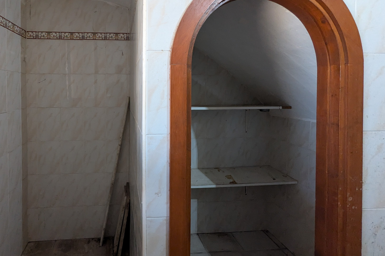 Under-stair storage nook featuring a wooden archway, white tiled walls with decorative borders, and built-in shelving.