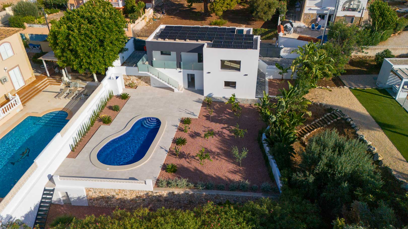 Aerial view of the modern villa featuring rooftop solar panels, a kidney-shaped pool, and a red marble gravel garden with natural stone walls.
