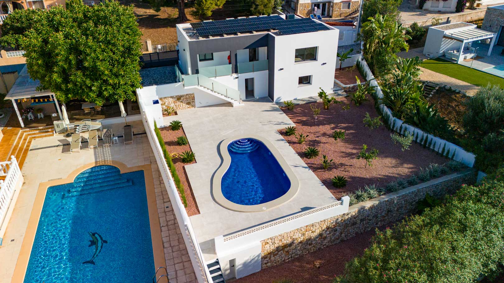 Aerial view of the modern villa featuring rooftop solar panels, an organic-shaped pool, red gravel garden, and natural stone walls. Glass railings accent the contemporary architecture.