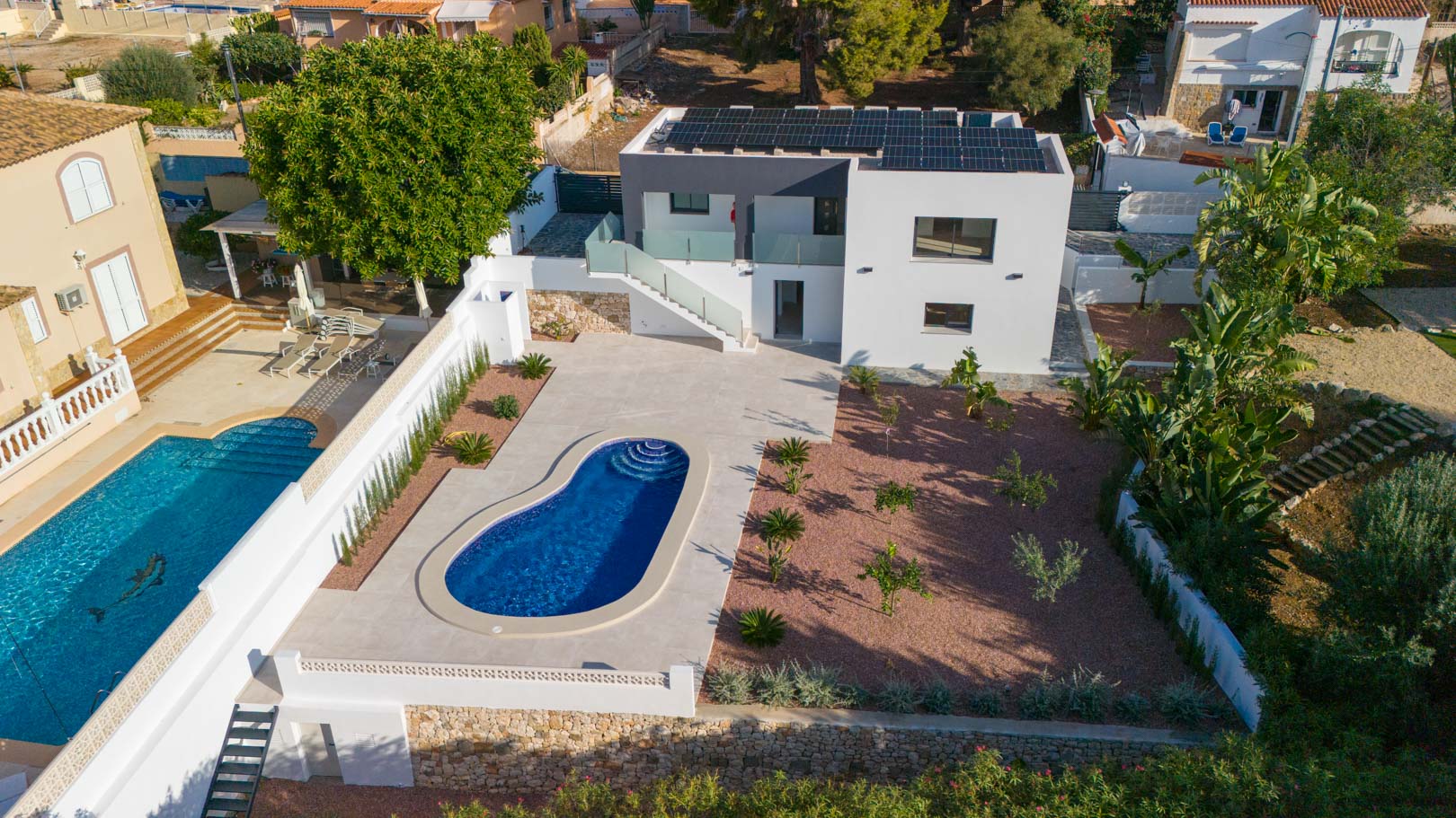 Aerial view of the villa featuring a kidney-shaped pool, red gravel garden, and solar panels on the roof. Includes glass railings and natural stone retaining walls.