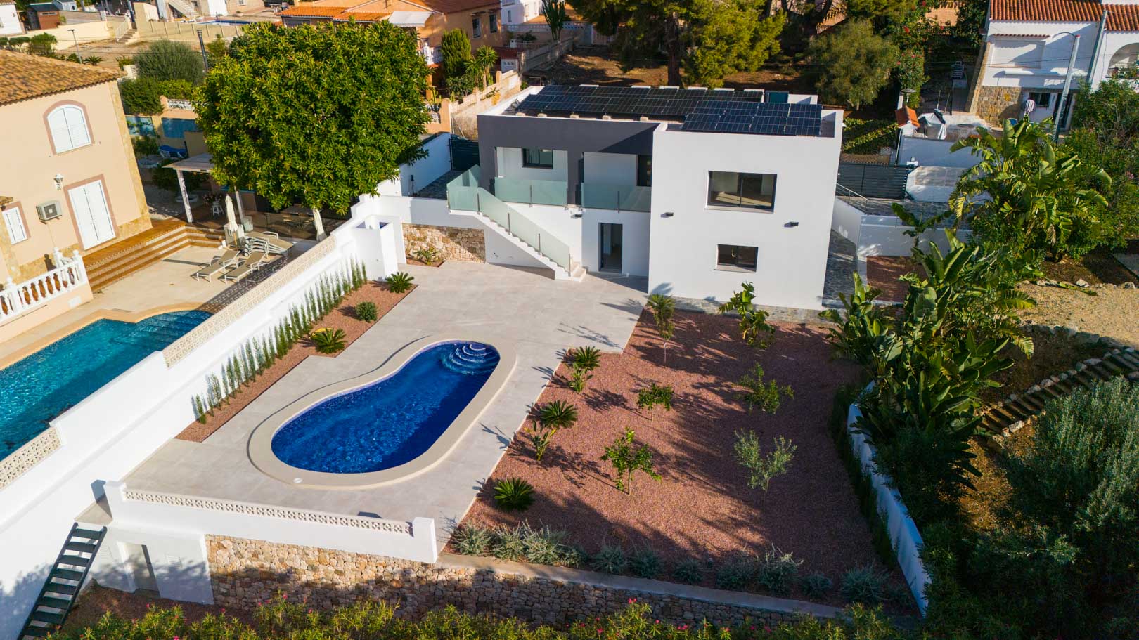 Aerial view showing the curved pool, red marble gravel garden, and rooftop solar panels. Features include glass railings and natural stone perimeter walls.