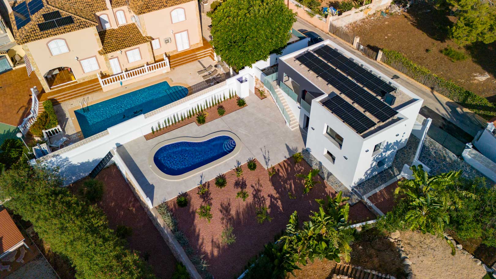 Aerial view showing the modern villa with rooftop solar panels, a kidney-shaped pool, and red marble gravel landscaping. Features glass railings and natural stone walls.