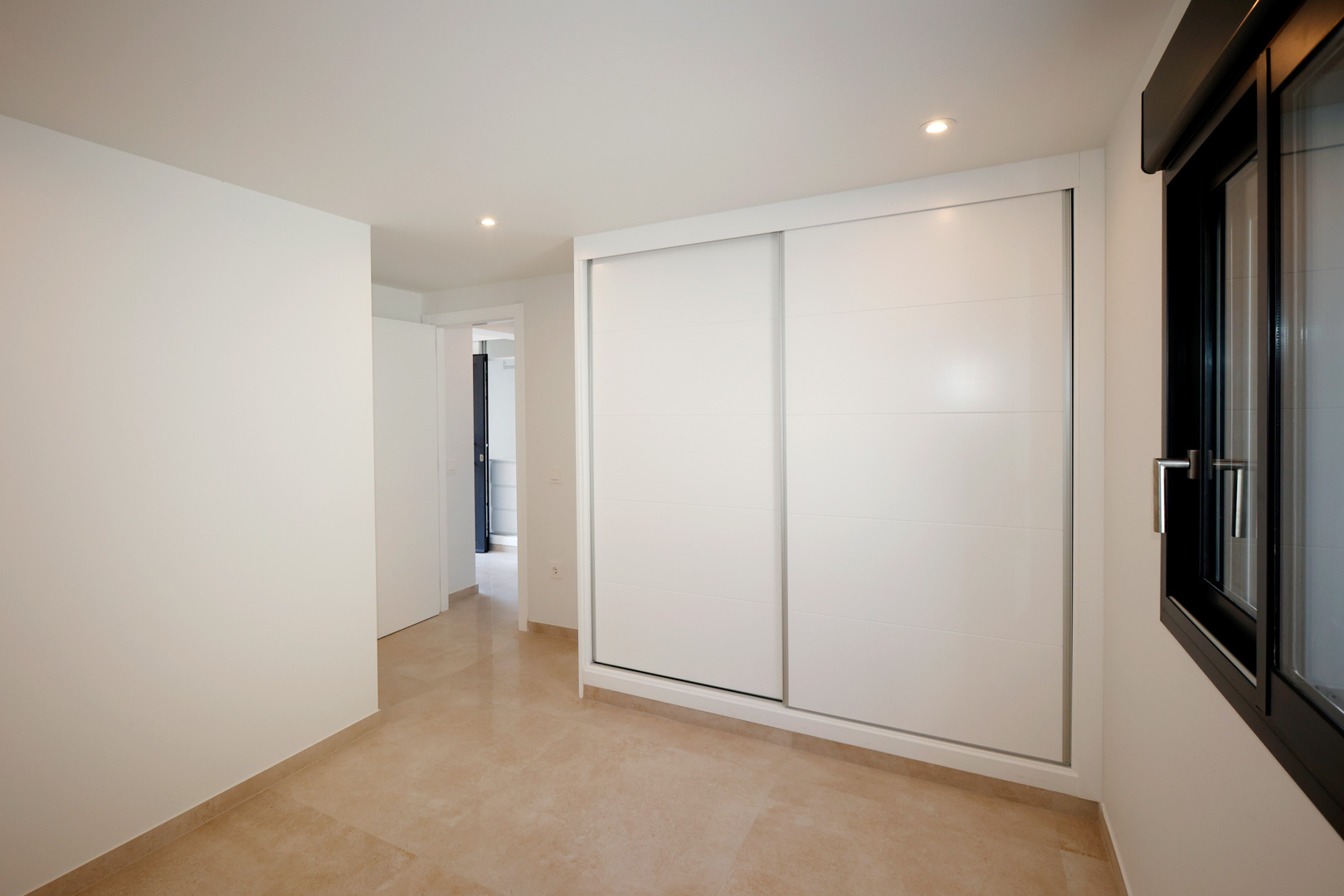Bedroom featuring a built-in wardrobe with white sliding doors, porcelain tile flooring, and recessed LED lighting next to a dark aluminum window frame.